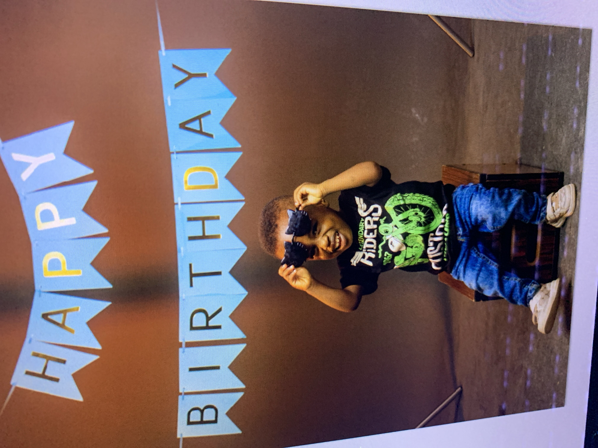 The image depicts a young child in a studio setting, celebrating a birthday. A blue banner with "HAPPY BIRTHDAY" written in gold and white letters hangs in the background. The child, wearing a black t-shirt with a green graphic and blue jeans, is seated on a wooden stool and is playfully holding a pair of novelty glasses over their eyes. The background is a plain brown backdrop, typical of a studio. There are no discernible elements in the image that would indicate a specific location in Gura Top, Nigeria.