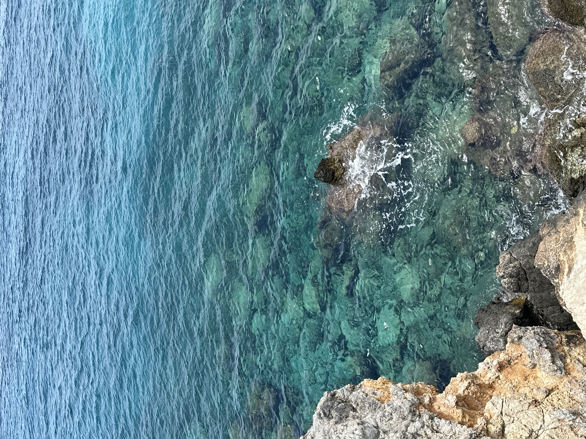 A high-angle shot captures the clear, turquoise waters of the Mediterranean Sea lapping against the rocky coastline of Rizokarpaso, Northern Cyprus. The water's transparency allows for a view of the submerged rocks and seabed, with gentle ripples disturbing the surface. In the foreground, rugged, textured rocks of varying shades of grey and orange form the edge of the land. The scene is devoid of any human presence or activity, presenting a natural and serene coastal landscape.