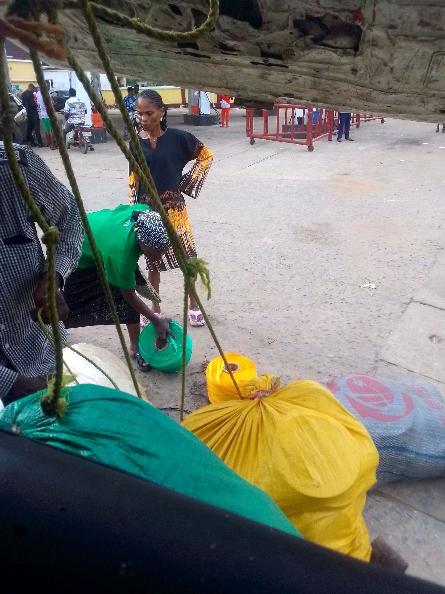 Ihiala, Nigeria: A woman in a green shirt and headscarf bends to examine a roll of green ribbon. Several brightly colored bags are on the ground next to her. A second woman stands with her hands on her hips, wearing a black and brown patterned dress. A man in a checkered shirt stands partially visible to the left. In the background, other people and a vehicle are present. A red metal barrier is also visible in the background. The view is partly obscured by ropes and a large, dirty, flat object overhead. The ground is paved.