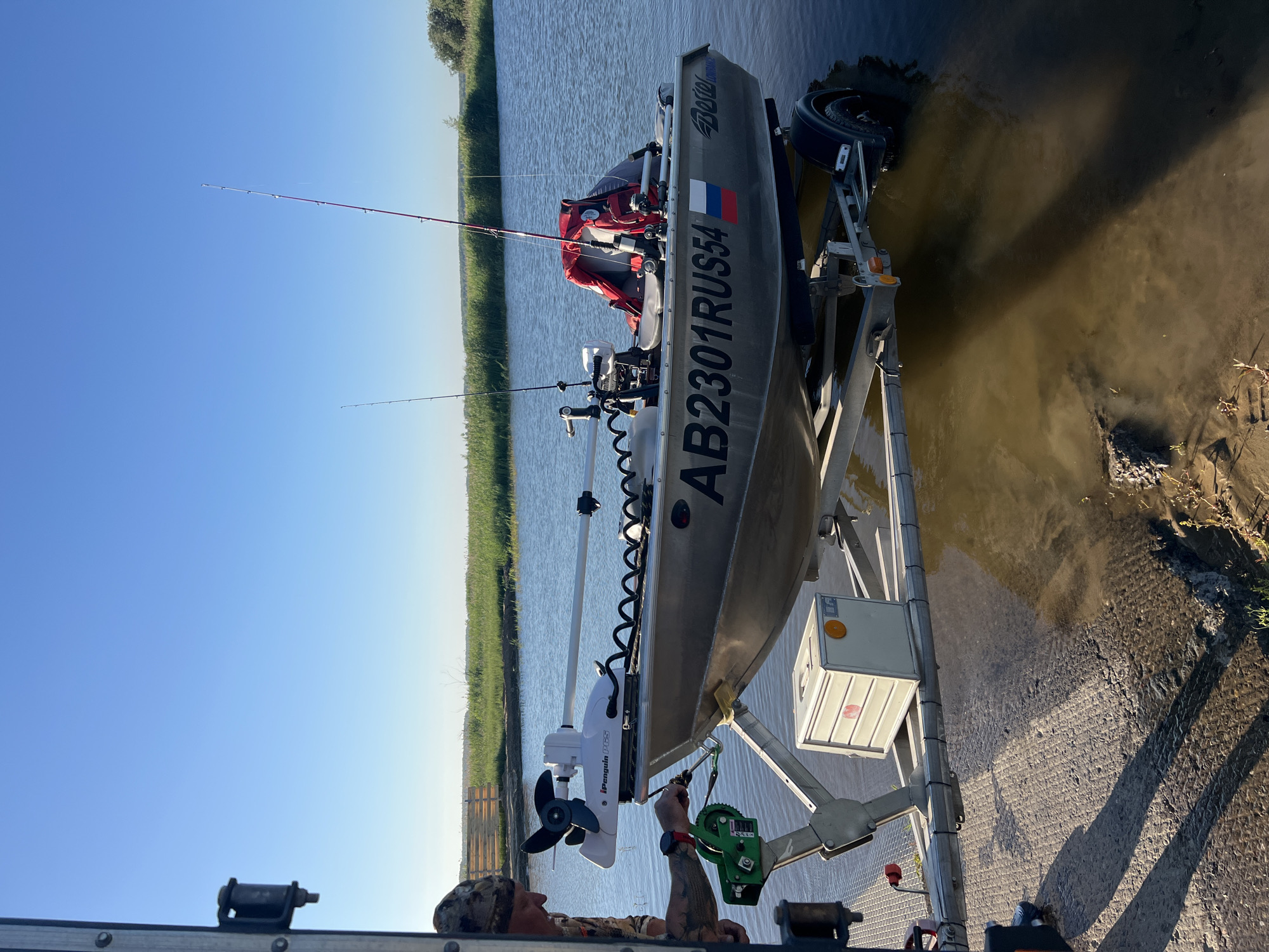 A man in camouflage clothing is preparing to launch a boat from a trailer into a body of water. The boat is silver and has the registration "AB2301RUS54" on its side, along with a Russian flag sticker. It is equipped with a trolling motor and two fishing rods. The trailer is partially submerged in the water, with one wheel visible. In the background, there is a green grassy embankment and a clear blue sky. The scene appears to be in the city of Novopichugovo, Russia.