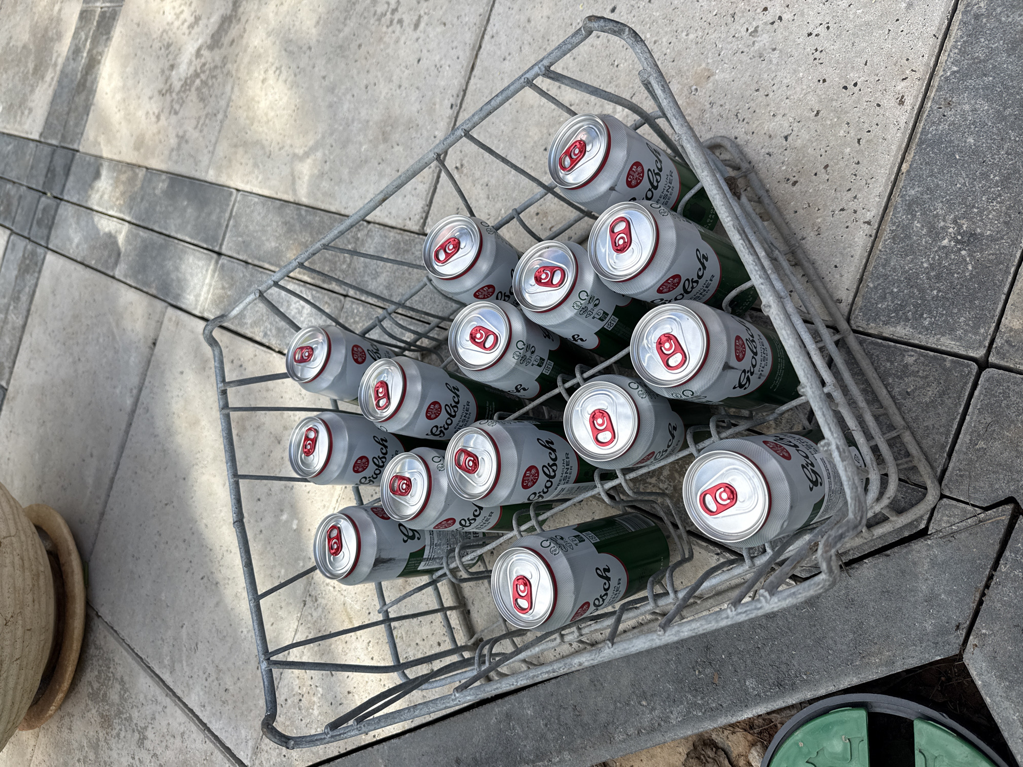 The image shows a metal wire basket filled with 15 cans of Grolsch Premium Pilsener beer. The cans are arranged in a neat grid pattern within the basket. The background consists of gray and tan paving stones, with a small portion of a large terracotta pot visible on the left and a green utility cover on the right. The scene appears to be outdoors. There is no evidence to indicate that this image was taken in the city of Гольяны, Russia.