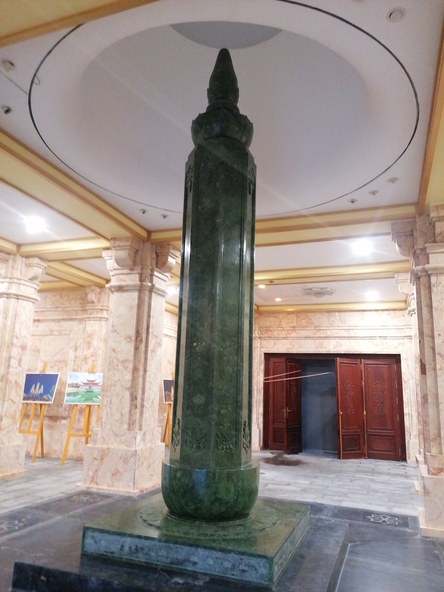 A tall, green, jade-like pillar stands prominently in a large, opulent room. The pillar has a bulbous base and a tapering top, with decorative carvings along its shaft. It sits on a multi-tiered platform of matching green stone. The room is characterized by marble columns, ornate ceiling details, and recessed lighting. Several paintings on easels are placed to the left. Double wooden doors, partially open, lead to another area. The scene is presumably taken in Hong Gai Ward, Vietnam.