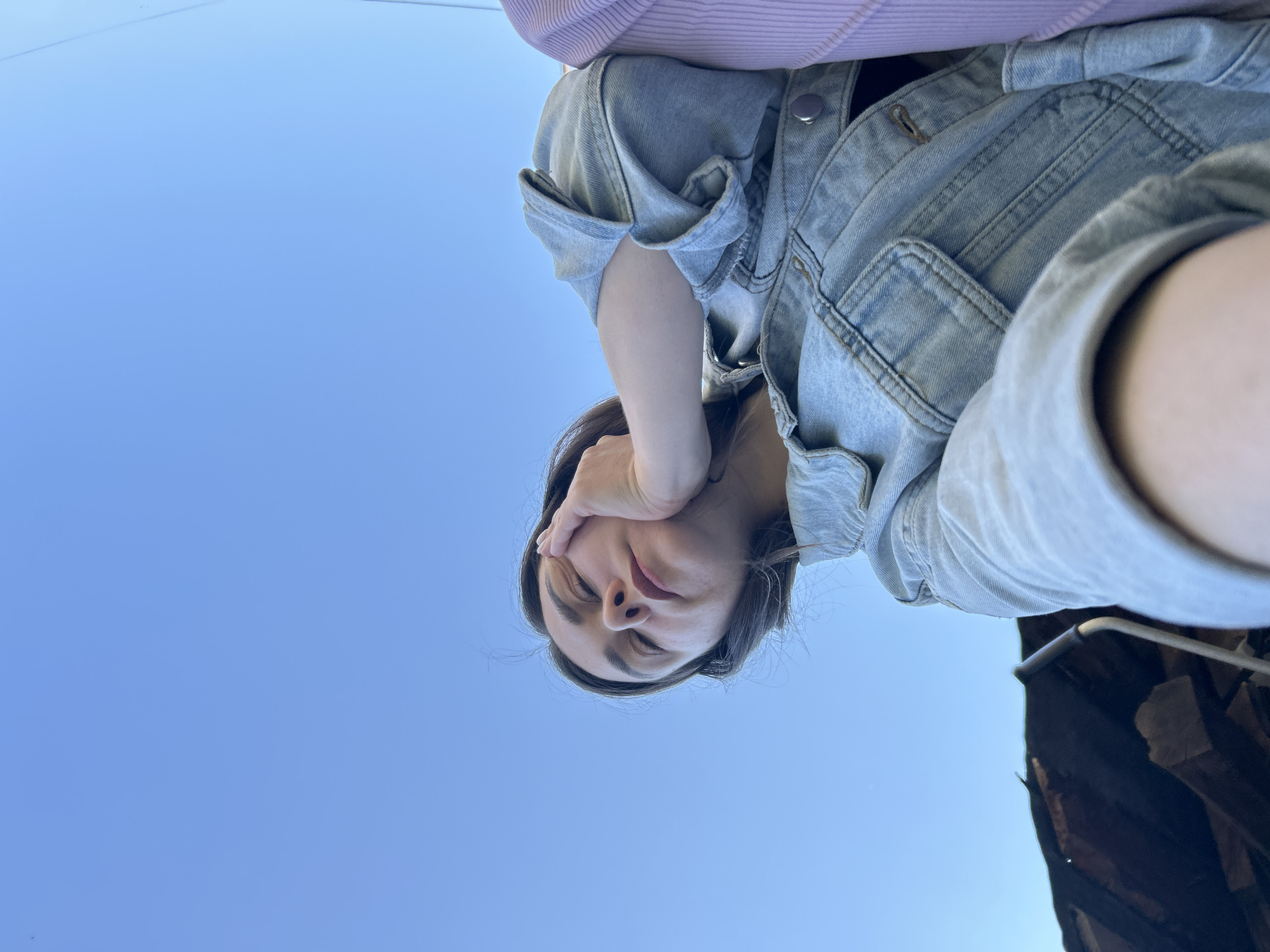The photograph is an upward-angled shot of a woman with her hand resting on her face. She wears a light-wash denim jacket and a lavender shirt. The background is a clear blue sky, except for the lower right corner, where a dark structure with possible wooden elements is visible. The photo was taken in Chaikovsky, Russia.