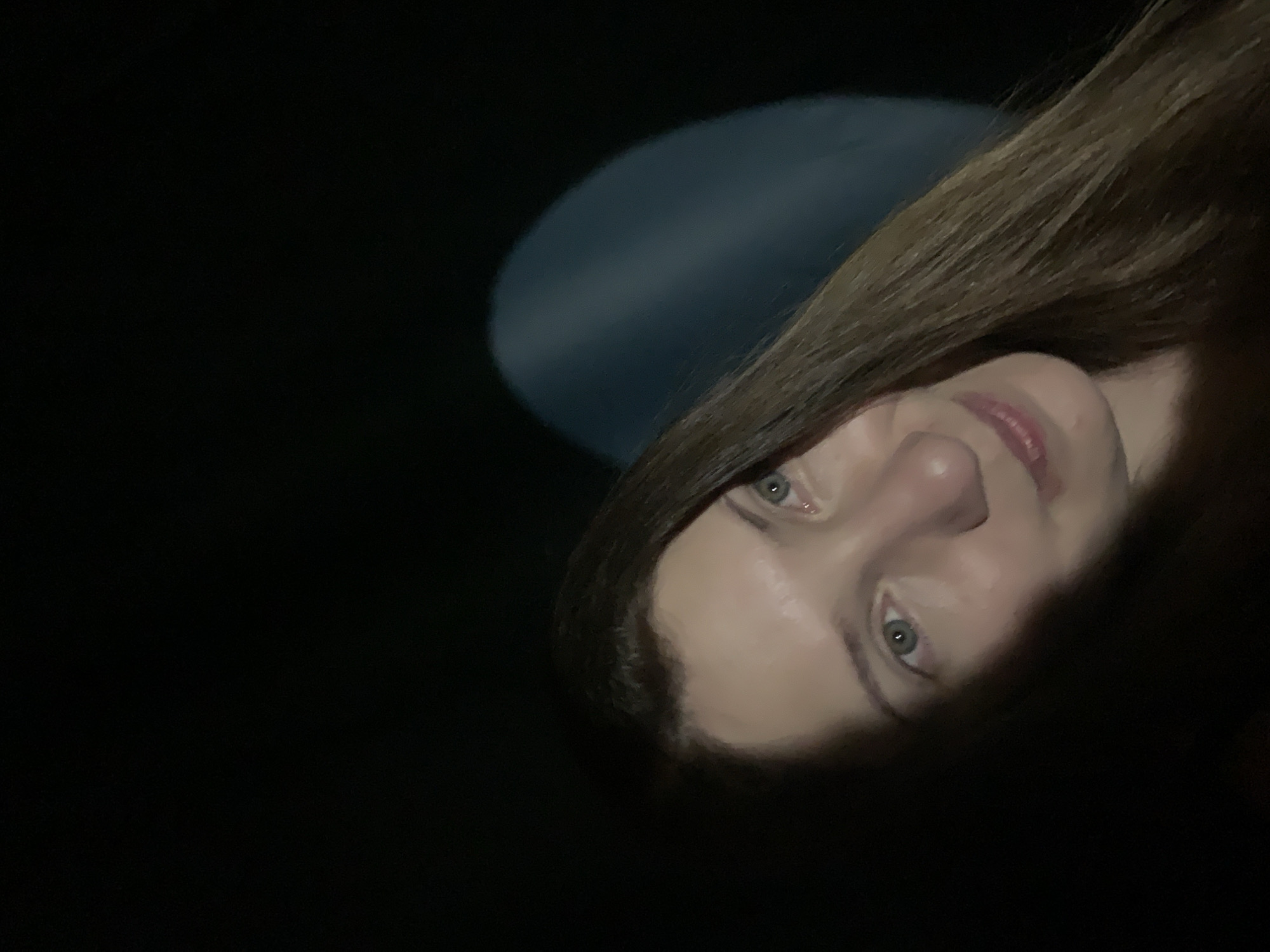 A close-up photograph shows a woman's face partially illuminated against a dark background. The woman has fair skin, light eyes, and brown hair. A blurred, circular shape of light is visible behind her. The location cannot be determined based solely on this image.