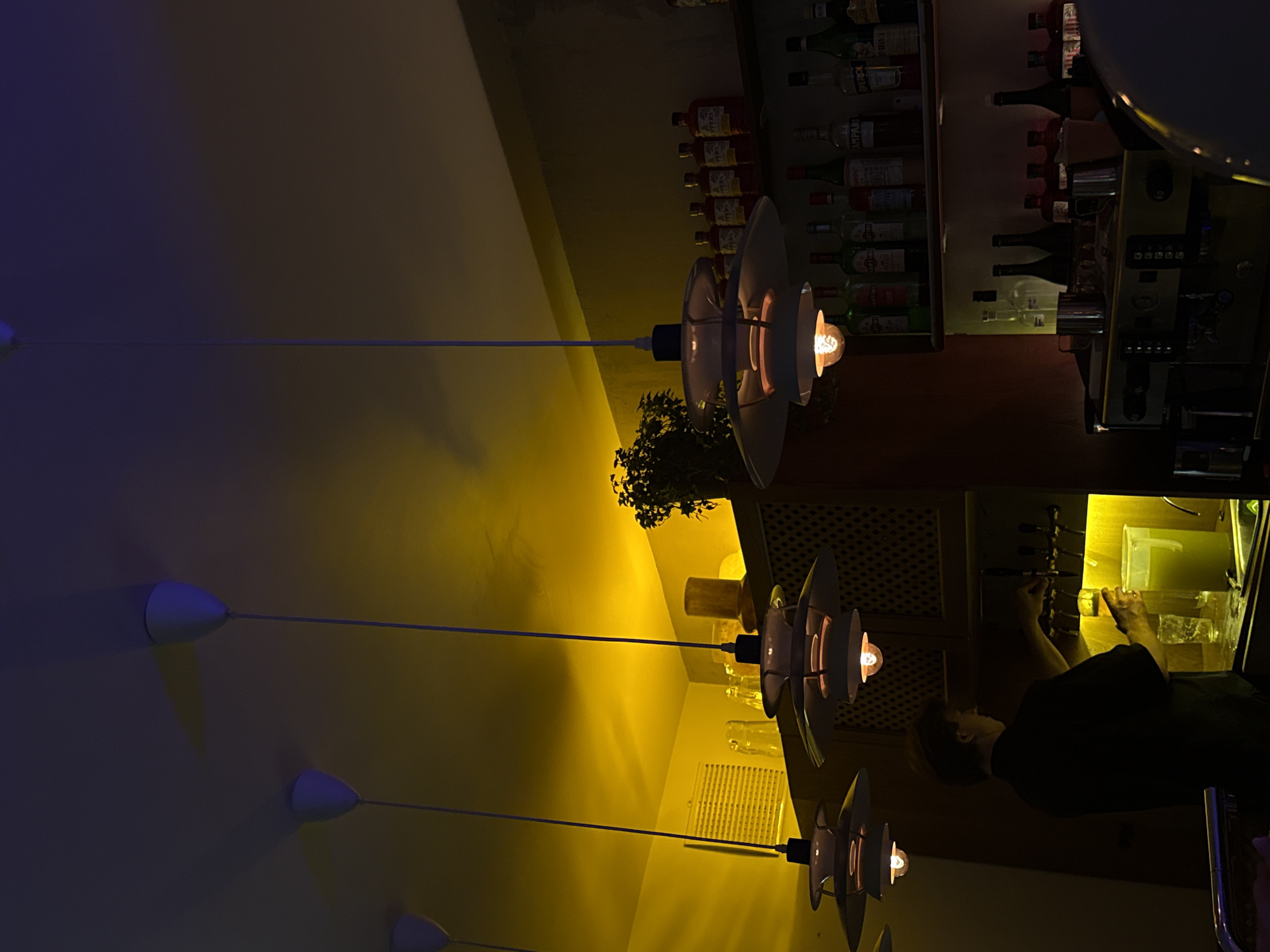 A rotated photograph taken indoors shows a bar setting. The lower portion of the image depicts a bartender serving a drink, and rows of liquor bottles are visible on shelves in the upper right corner. Three multi-tiered lamps with lit bulbs hang from the ceiling, creating a warm glow. The upper left side of the image fades into a darker blue-grey color. No identifying information explicitly places the scene in Krasnoyarsk, Russia.