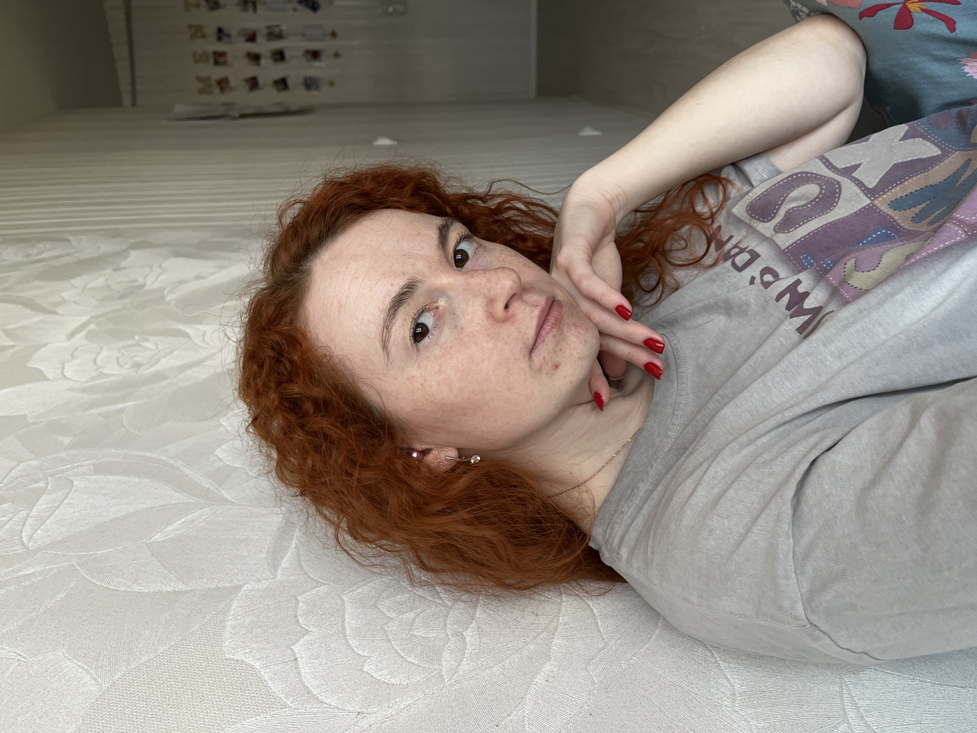 A woman with red curly hair and freckles is lying on a white mattress. She is wearing a gray t-shirt and has red nail polish. Her left hand is near her face. The location is likely Chaikovsky, Russia. In the background, out of focus, are a wall, trim, and several photos.