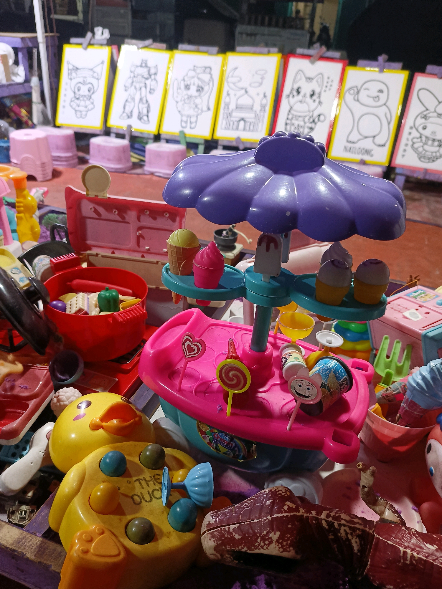 This image depicts a collection of colorful children's toys, possibly for sale or display at an outdoor market in Tangerang, Indonesia. In the background, several white boards with yellow frames are propped up on easels, displaying line drawings of cartoon characters, robots, and a mosque. Arranged in front of these drawings are small pink plastic stools.
The foreground is dominated by various toy items. Prominently featured is a pink and purple toy ice cream stand with miniature ice cream cones and popsicles. To the left, a red toy grill is filled with toy food items including a hot dog, a green bell pepper, and what appears to be bread. A bright yellow duck toy with large eyes and an open beak is visible, with a blue propeller-like object attached to its back, and the words "THE DUCK" printed on its belly. Other scattered toys include a pink suitcase, a small yellow bottle with an orange top, and parts of what might be a toy kitchen set. The overall impression is one of a vibrant, playful assortment of items, possibly set up at a night market or a temporary stall. The lighting suggests it is evening or nighttime.