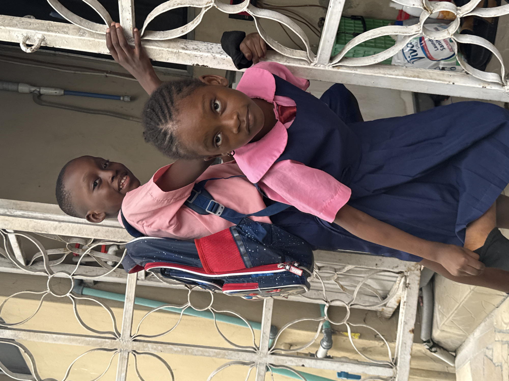 Two children, a boy and a girl, stand behind a white, metal security gate. Both wear school uniforms with pink shirts; the girl also wears a navy blue jumper dress. The boy has a backpack. The girl is taller and leans over the boy. They both look towards the camera. The background includes a light yellow wall and exterior pipes. Ado, Nigeria, is a likely location given the style of security gate and school uniforms.