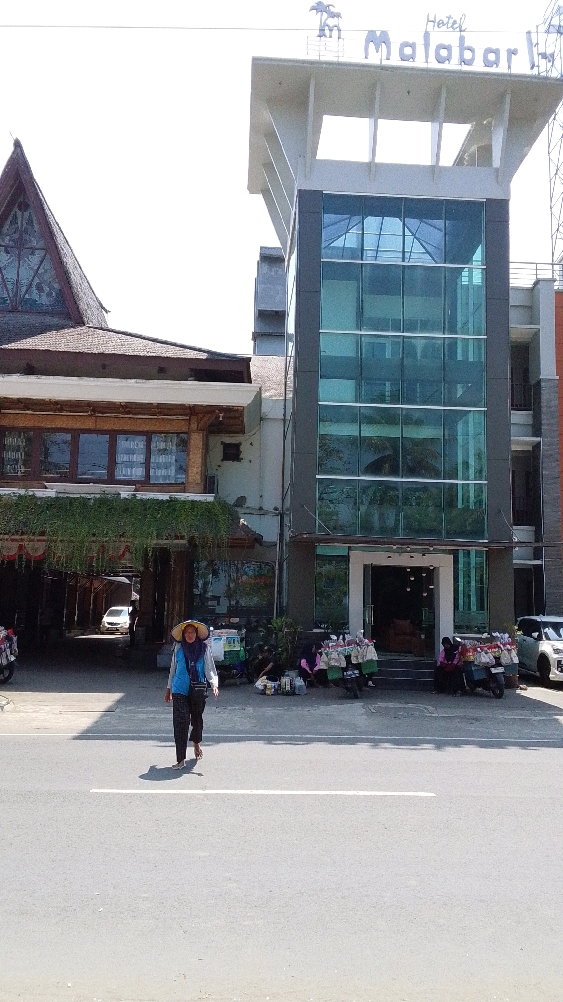 The image depicts a street scene in front of the Hotel Malabar in Desa Pagandaran, Indonesia. A person wearing a straw hat, a blue vest, and checkered pants is walking on the asphalt road. In the background, there is a building with a modern glass facade and a sign that reads "Hotel Malabar." To the left of the hotel, there is another building with a dark tiled roof and wooden accents. Several motorcycles are parked in front of the hotel, some with vendors selling goods. A white car is visible on the far right of the image, and a white car is parked in the archway of the building on the left. The sky is bright and clear.