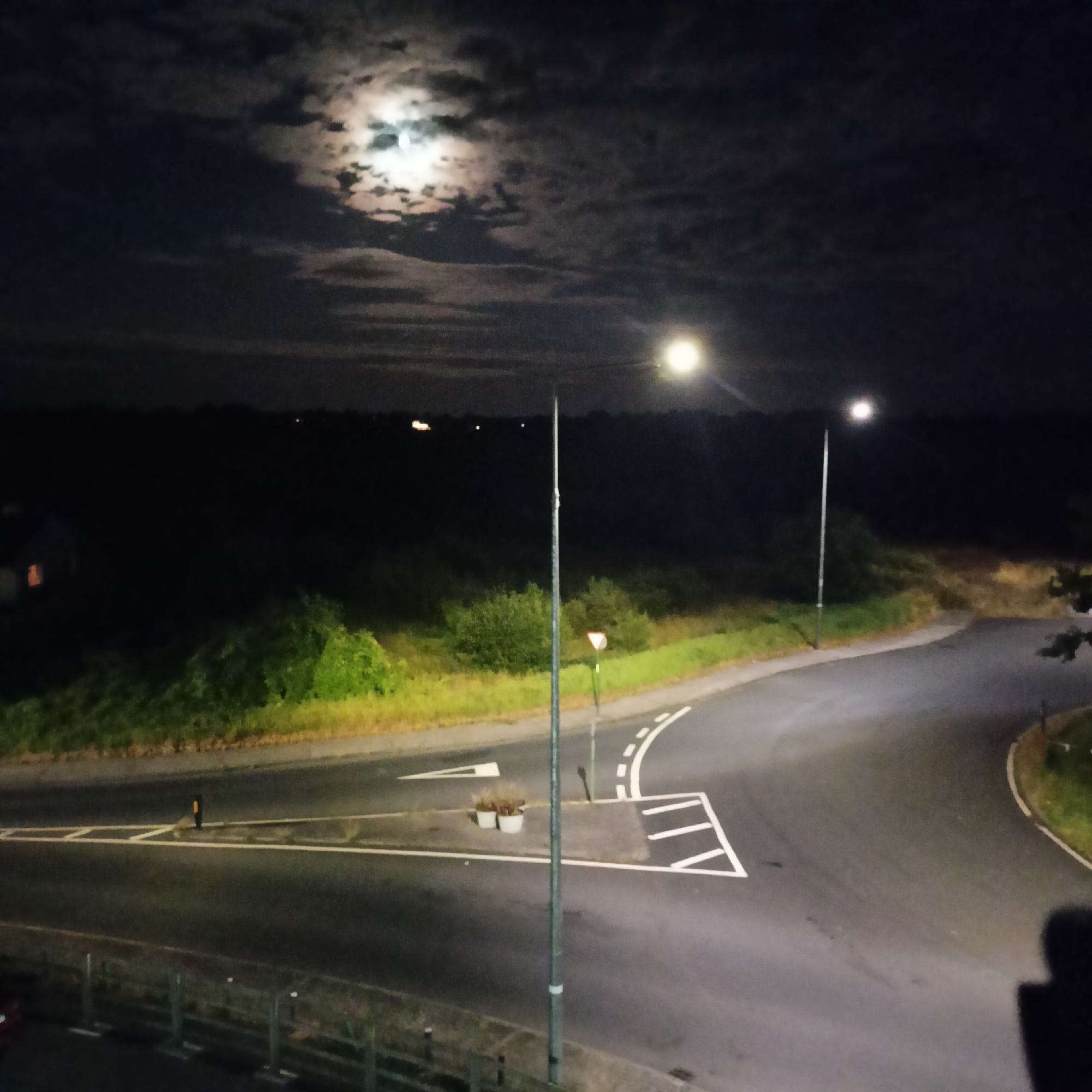 The image captures a night scene in Limerick, Ireland, featuring a moonlit sky with scattered clouds. In the foreground, a curved road with white line markings is visible, illuminated by streetlights. A tall lamppost stands centrally, with another to the right. Beyond the road, dense green foliage lines the landscape, and in the distance, faint lights suggest the presence of buildings or other structures. To the left, a portion of a building with a lit window is discernable. The overall atmosphere is dark and tranquil.