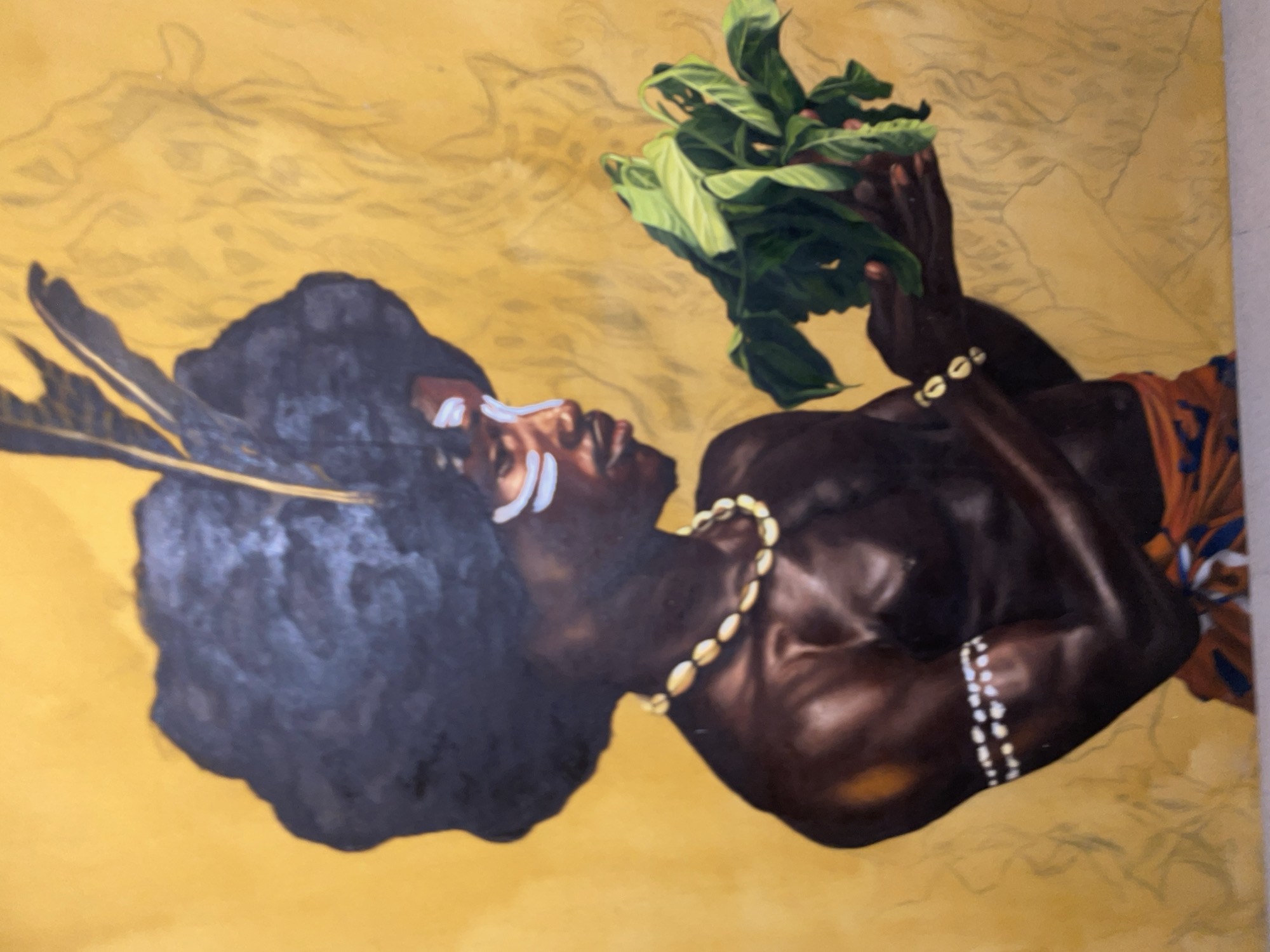 The image is a painting of a Black man, likely of Nigerian descent, holding a bundle of green leaves. He lies on his side against a yellow background. He wears what appears to be white ritualistic paint on his face, feathers in his hair, and a beaded necklace. He also wears a beaded bracelet. His skin is painted with highlights to accentuate his musculature. He is partially covered by a garment with an orange and blue pattern. The style of the painting suggests a traditional, possibly indigenous, Nigerian cultural context. It is not possible to determine its specific location within the city of Ado, Nigeria, from the visual information given. There are no other people visible in the scene, and the image primarily depicts a single, static subject. There is no discernible interaction beyond the man holding the leaves. The environment is plain and seems artistic rather than representative of a specific location.