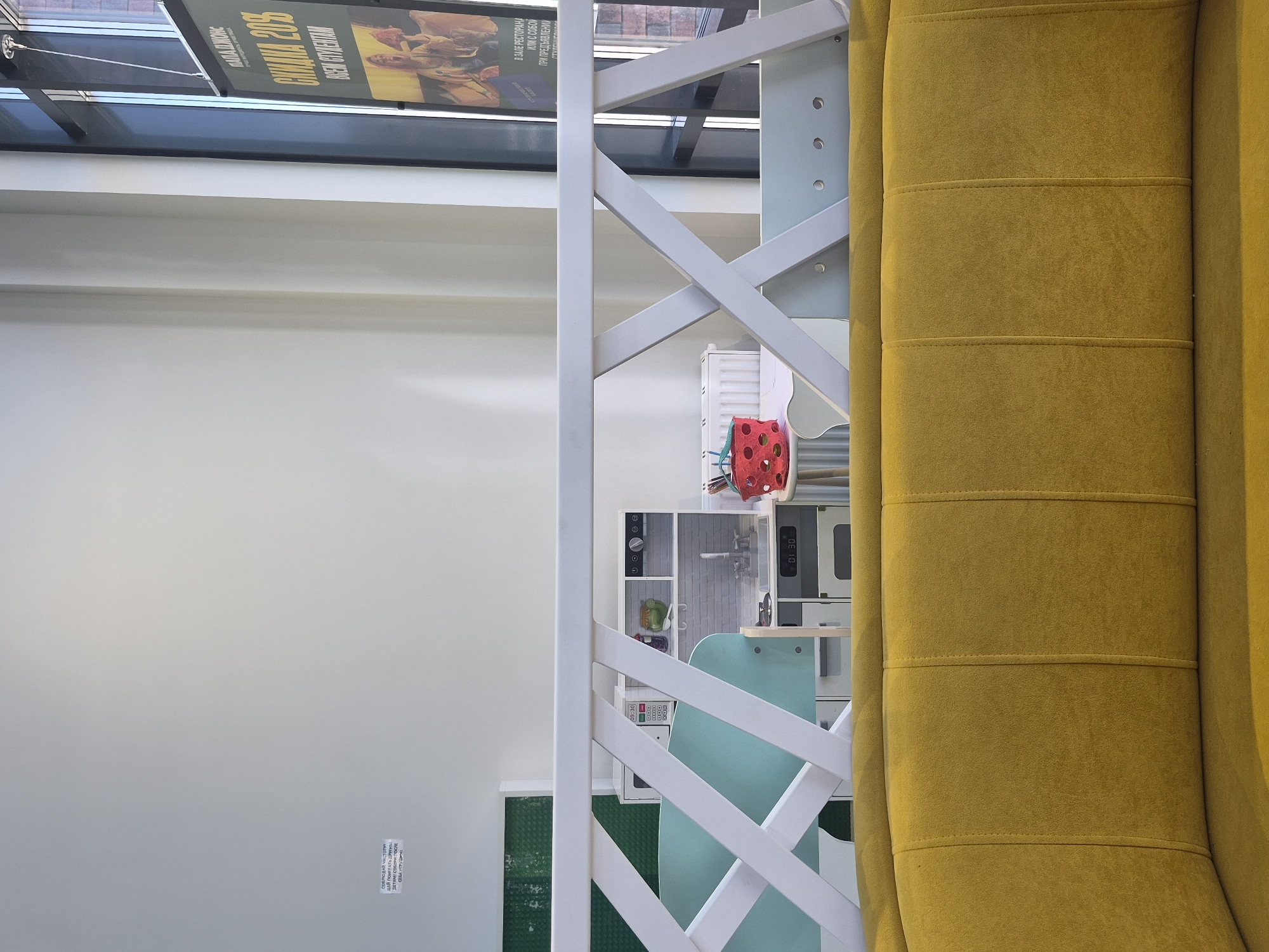 The image shows a room in what appears to be a child's play area. The dominant elements include a mustard-yellow upholstered object (possibly a piece of furniture like a sofa or chair), a white wooden structure with criss-cross bars, and a play kitchen. The play kitchen features various toy appliances and utensils. In the background, there is a poster with the name "Пала Джонс" and the phrase "В ЗАЛЕ РЕСТОРАНА ИЛИ С СОБОЙ ПРИ ПРЕДЪЯВЛЕНИЙ," as well as the time "09:30." The poster is advertising something and includes the text "СОБЛЮДАЙ Чистоту," "ДАЙ ПОИГРАТЬ ДРУГИ," and "ДЕТЯМІ СОБЕРИ ПОСЛЕ." The presence of Cyrillic text suggests the location is in a Russian-speaking region, possibly Russia. Based on other information associated with the image, it is likely in Izhevsk, Russia.