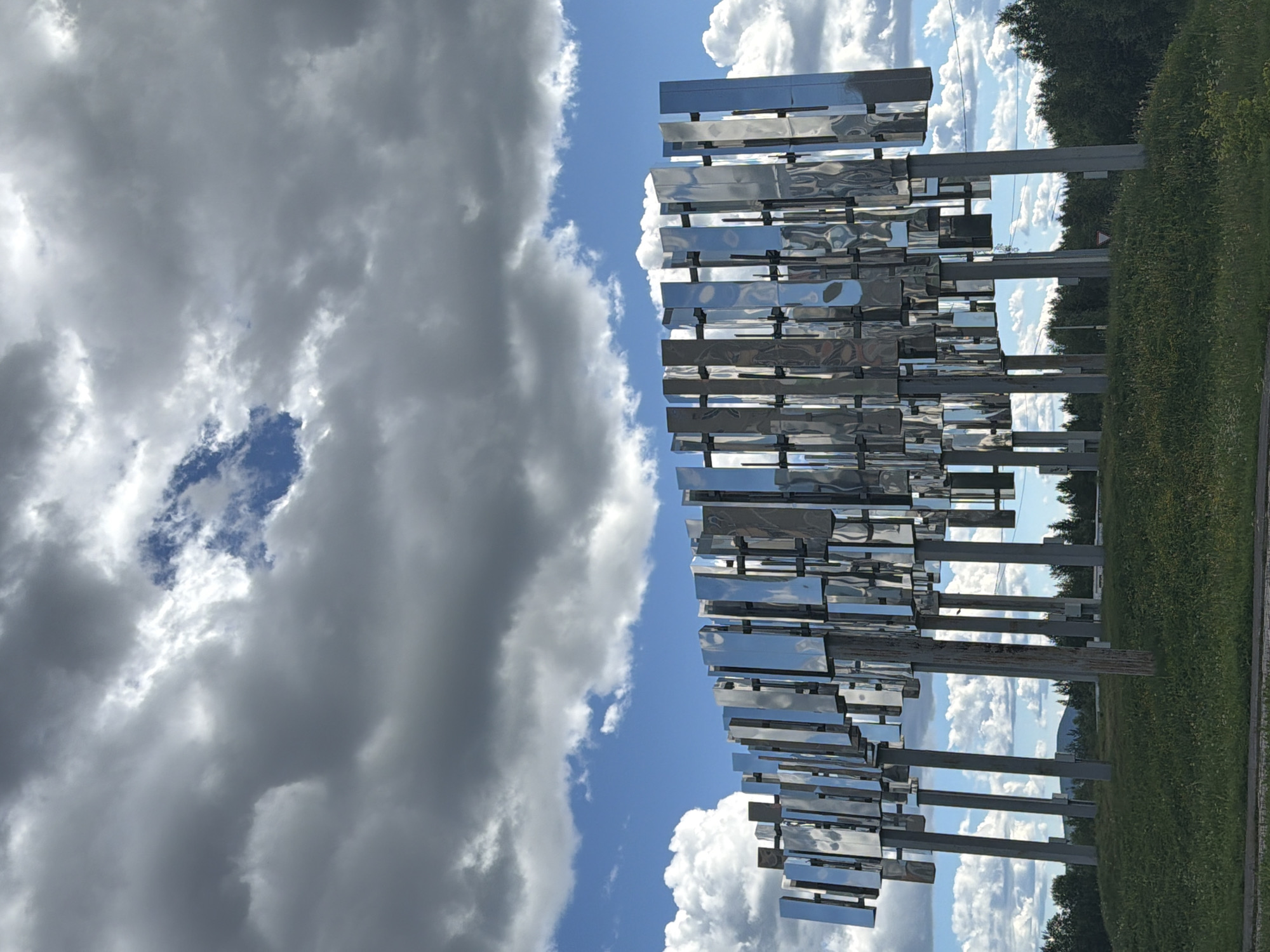 A reflective sculpture composed of numerous vertical, metallic rectangles stands on a grassy hill under a sky filled with dramatic, grey and white clouds interspersed with patches of blue sky. The sculpture is supported by a series of grey poles. In the background, a dense line of green trees forms the horizon, with a distant, hazy mountain visible on the left. The foreground is a sloping green lawn with some visible white and yellow wildflowers. This sculpture is located in the city of Polarnie Zori, Russia.
