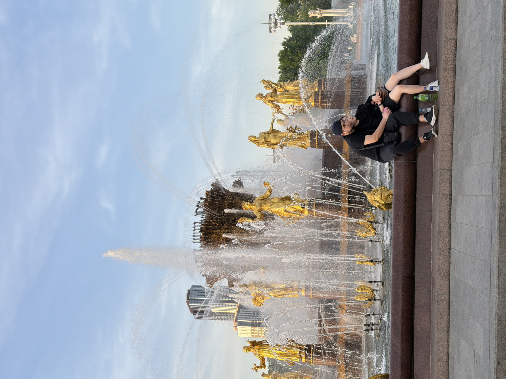 A couple is sitting at the edge of the Friendship of Nations Fountain in Moscow, Russia. The fountain features numerous gold statues and water spouts. A man in a black shirt and pants, and a baseball cap is seated with a woman. The woman has bare legs and is wearing shorts and sneakers. Water bottles are sitting near them on the platform. The fountain is set against a backdrop of a blue sky with some clouds. Buildings are visible behind the fountain.