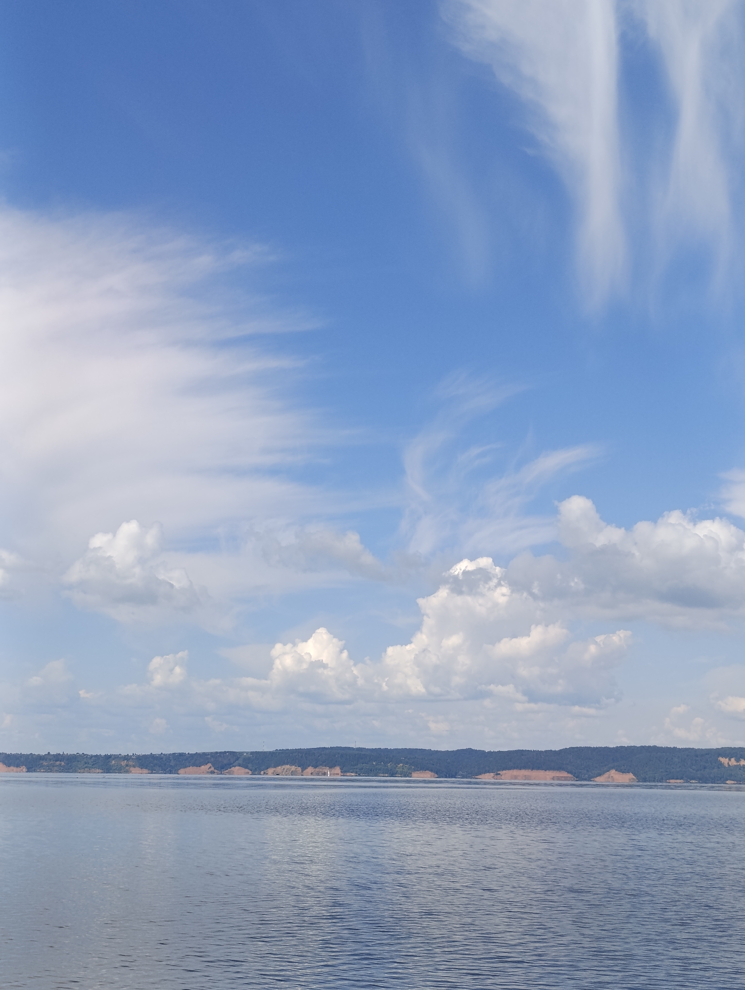 The image depicts a wide, calm body of water under a blue sky with scattered white clouds. The horizon line is low, with a distant, wooded shoreline marked by sandy bluffs. A small white sailboat is visible on the water near the shoreline. The scene appears to be a natural landscape, possibly a river or lake. The location is identified as Vekoshinka, Russia. No people or specific actions are discernible in the image.