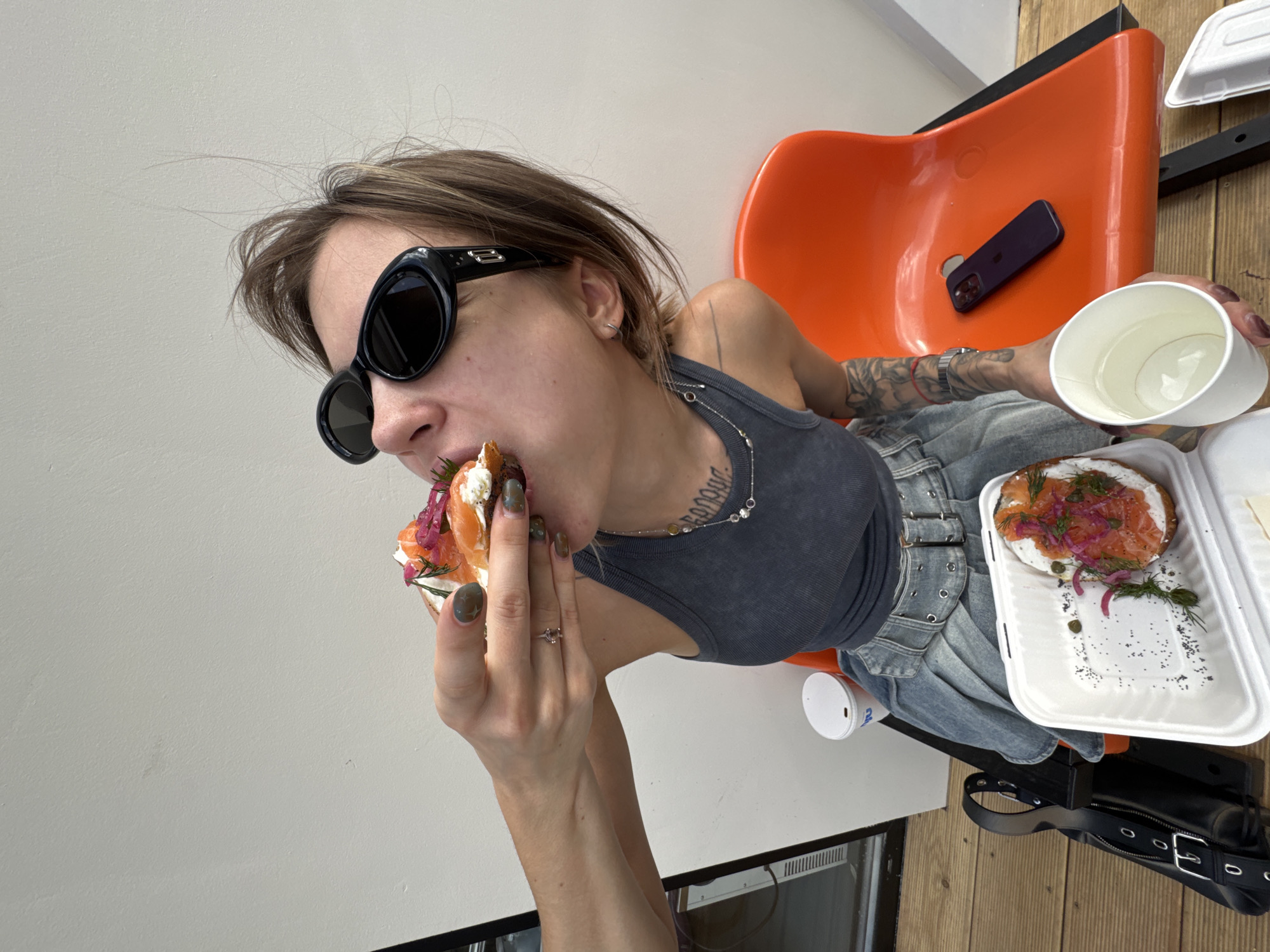 A woman with short brown hair and sunglasses is eating a sandwich with salmon, cream cheese, dill, and red onions. She is sitting in an orange chair with a black smartphone resting on it. She is wearing a gray tank top and light-wash denim pants with grommets and buckles. Her left hand is holding the sandwich, and her right hand is holding a white cup. A white takeout container with a salmon bagel, a white cup, and a black purse are visible on the wooden floor. A white wall is to the left of the chair. The location is unknown, and there is no information to confirm it is in Moscow, Russia.