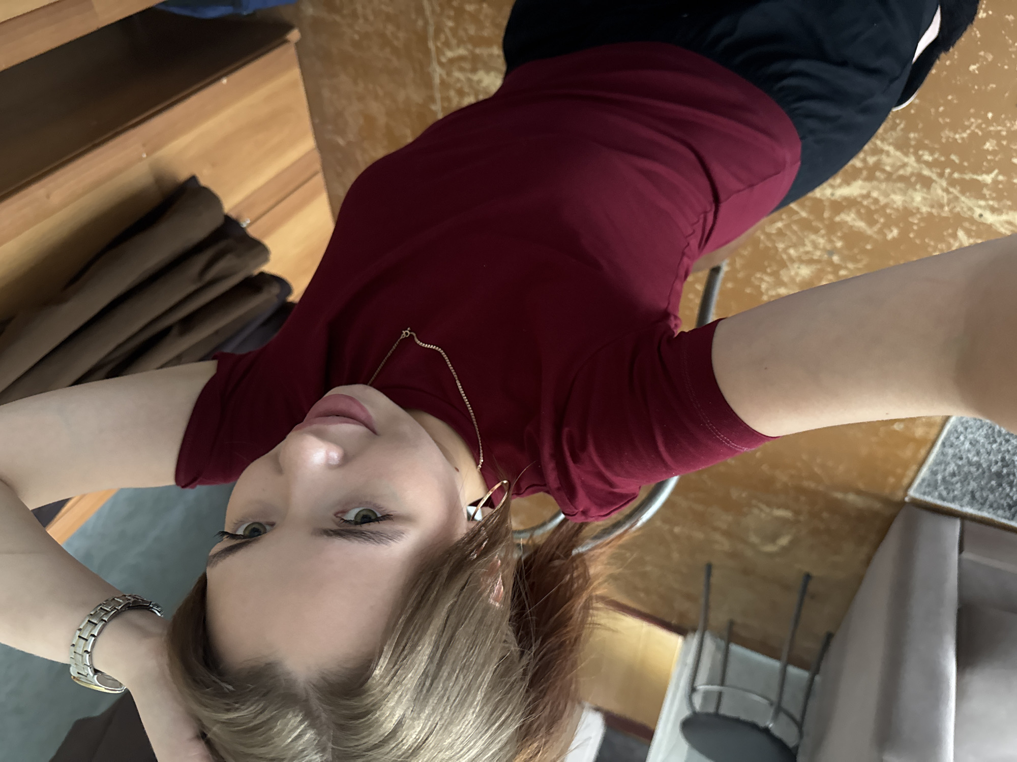 This is a first-person, eye-level, medium shot of a young woman with light skin and blonde hair. She is looking directly at the camera, with her hands behind her head. She is wearing a maroon t-shirt and dark pants, and a silver watch on her left wrist. She has light green eyes and is wearing subtle makeup, including mascara and lipstick. The background is blurred, but appears to be an indoor setting with wooden furniture and flooring. There are hints of a rug and a sofa in the background. The lighting suggests it could be daytime. The woman's expression is neutral, and the overall mood is casual and relaxed. There is no visible text in the image.