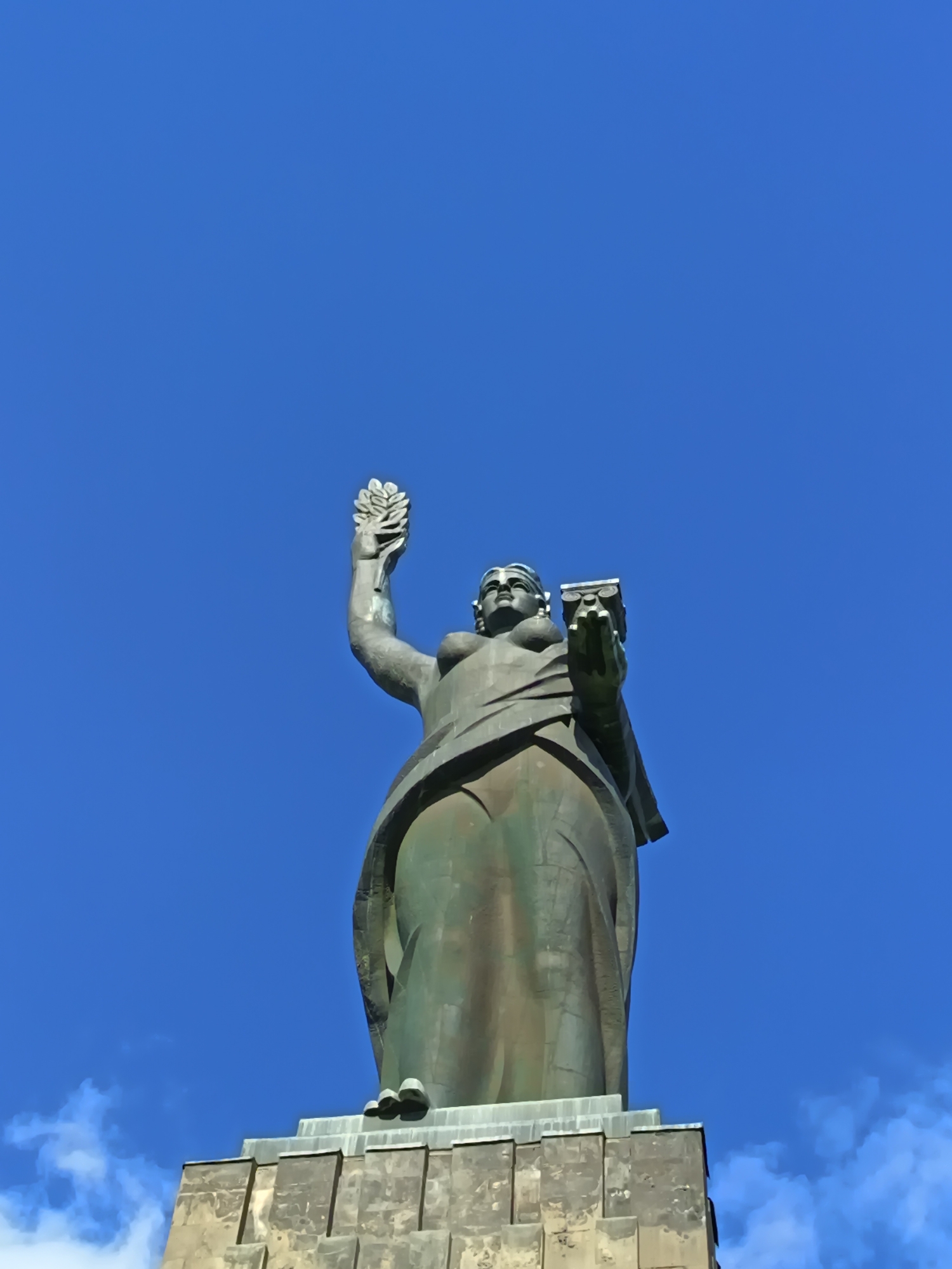 A low-angle, vertical photograph depicts the towering "Mother Armenia" statue against a clear blue sky with some wispy white clouds at the bottom left. The statue is mounted on a stone pedestal with a tiered, blocky design. The statue itself is of a draped female figure, likely made of bronze or a similar metal, giving it a green-brown hue. The figure holds a laurel branch in her raised right hand and a tablet or scroll in her left hand. The statue appears to be located in a city in Unknown, Unknown.