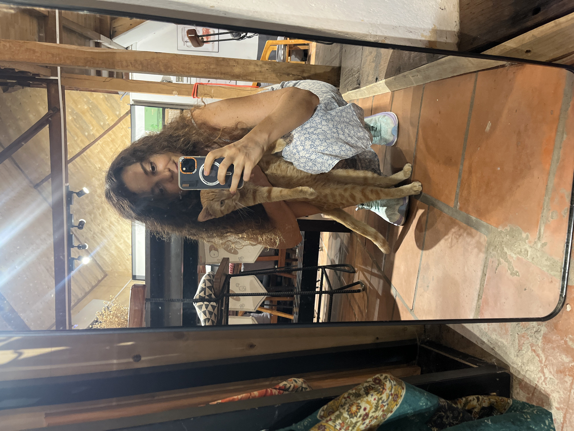 A young woman with curly brown hair is taking a mirror selfie while crouching and holding an orange tabby cat. She is wearing a light blue dress with a floral pattern and light blue sneakers. The cat is held in her left arm and looking towards the camera. The background appears to be an indoor space with exposed wooden beams on the ceiling and terracotta tiled flooring. In the reflection, there are shelves with items and some chairs. The scene is lit by overhead lights. The location is identified as Phu Thuy Ward, Vietnam.