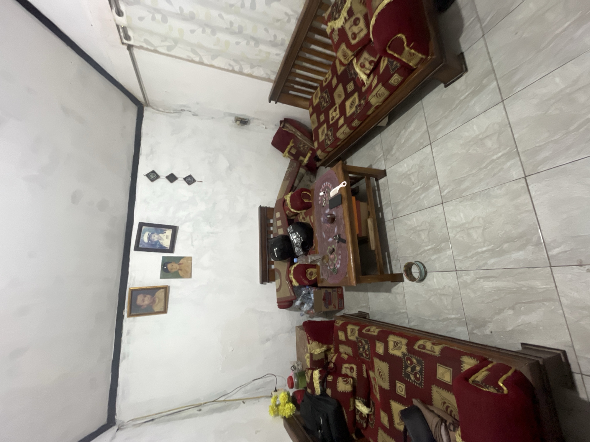 The image depicts a room in Pekalongan, Indonesia, likely a living room, furnished with several wooden seating units covered in red and gold patterned fabric. The room has white walls with some water stains and cracks, and a white ceiling with a black border along the eaves. On the wall, there are three framed pictures of children and two diamond-shaped decorative items.
In the center of the room is a wooden coffee table with a few items on it, including a phone, a comb, and some drinks. Behind the coffee table is a wooden seating unit with two helmets placed on top, one black and one black and white. To the left of the coffee table, there is another wooden seating unit, and a small table with a vase of yellow flowers and a red jar. To the right of the coffee table, a wooden sofa with similar red and gold patterned cushions is visible. The floor is tiled with light gray and white marbled tiles. The overall impression is of a simple, lived-in space.