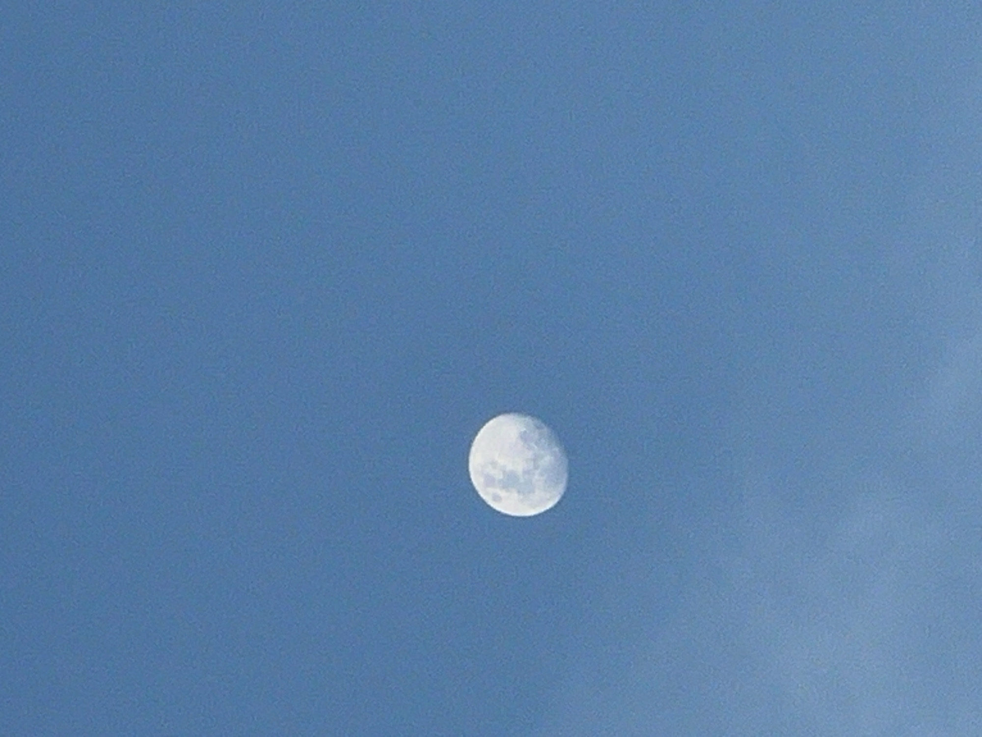 A daytime view of the moon in the sky, captured in Canggu, Indonesia. The moon, appearing almost full, is positioned in the center of the frame against a clear, blue sky. Faint clouds are visible in the lower-right corner of the image. No people or man-made objects are depicted in the scene.