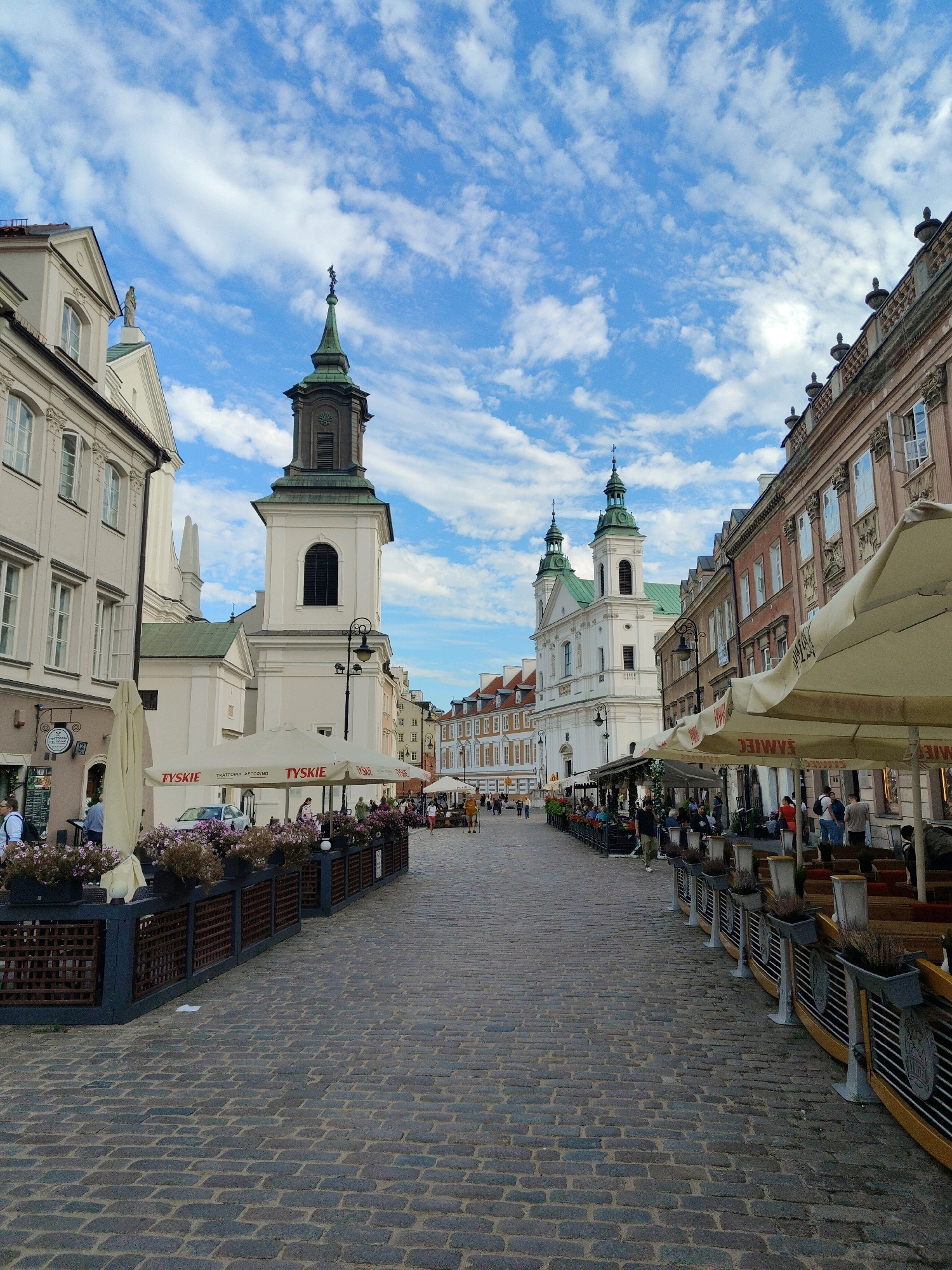 The image depicts a street scene in Warsaw, Poland, likely the Old Town. The perspective is from a cobblestone street looking down a narrow, paved lane lined with historic buildings and outdoor seating areas for cafes and restaurants. On the left, a light-colored building with multiple windows and arched details is visible, followed by a white church with a dark spire. To the right, the street is bordered by multi-story buildings with reddish-brown facades and numerous windows. Outdoor umbrellas, some bearing the "TYSKIE" logo, provide shade over tables and seating. Several people are present, appearing as patrons of the outdoor establishments or pedestrians. The sky above is blue with scattered white clouds. The overall atmosphere suggests a vibrant and historic urban setting.