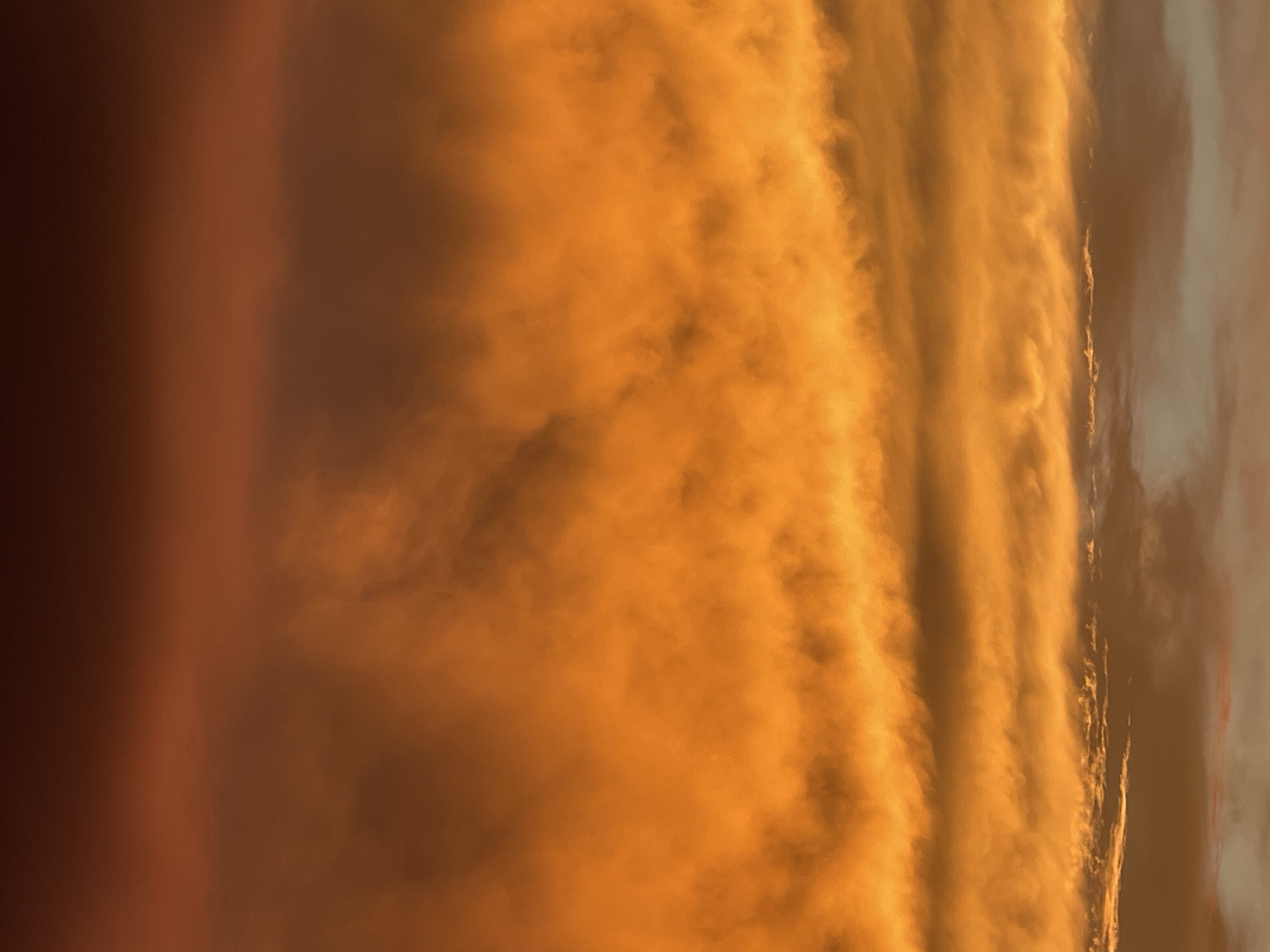 The image displays a dramatic sky filled with vibrant, fiery orange and deep red clouds, suggesting either sunrise or sunset. The clouds are textured and layered, with some appearing wispy and others more dense. The colors range from bright, almost golden orange in the lower and middle sections to darker, reddish-brown hues towards the top. The lighting is intense, illuminating the undersides of the clouds and creating a sense of depth. There are no discernible landmarks, people, or objects visible, making it a purely atmospheric scene. The location in Bluewater, Canada, is not visually represented in the image itself.