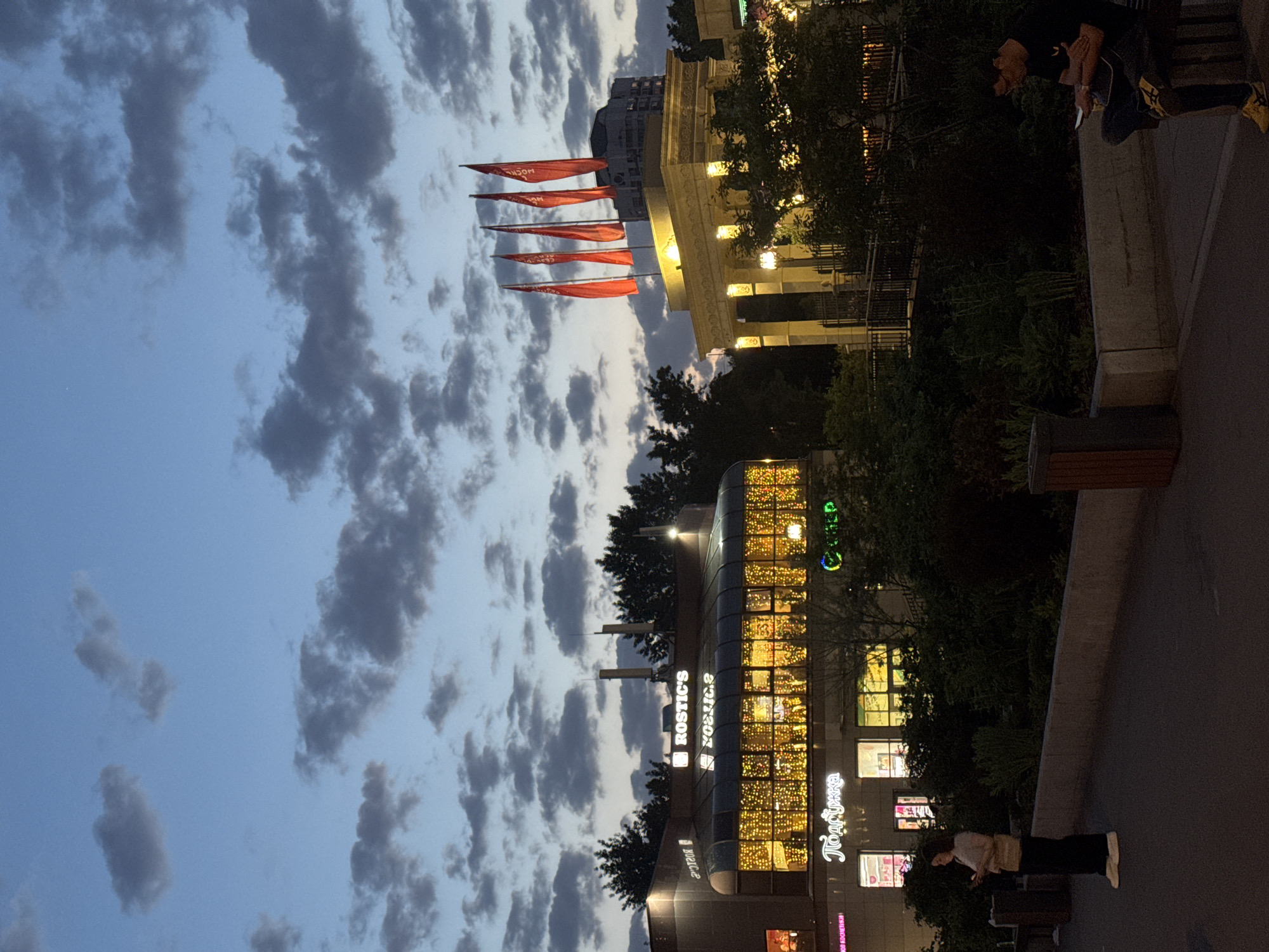The image shows an evening scene in Moscow, Russia. The sky is a cloudy blue with numerous grey clouds. In the distance, there is a row of red flags. A building with lit windows is visible, featuring "Rostic's" and other store signs in Cyrillic. There are people on the pavement, including one looking at a phone. Trees are interspersed with the buildings. The architecture includes classic pillars and modern building designs.