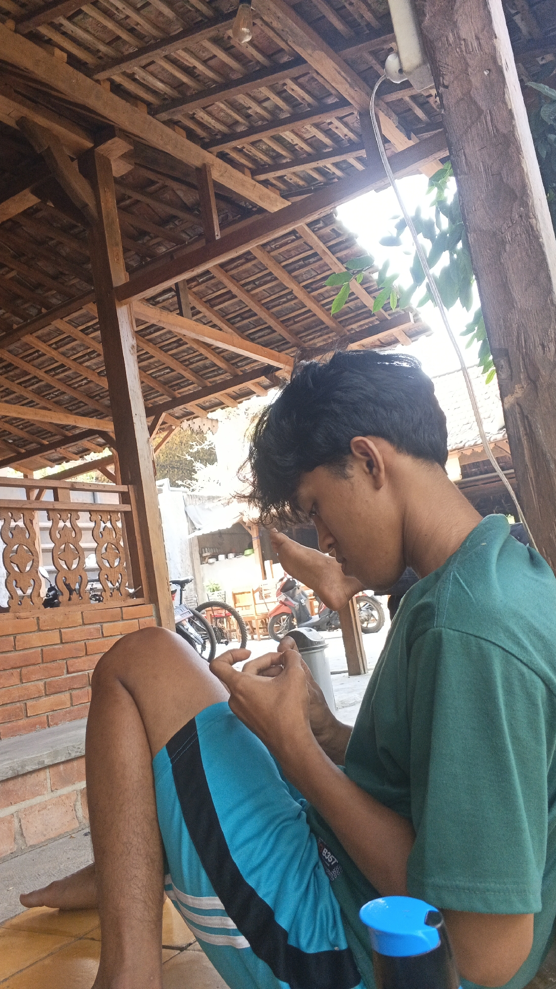 A young male, appearing to be of Indonesian ethnicity and in his late teens or early twenties, is seated outdoors under a wooden roof structure, possibly a pavilion or veranda, characteristic of traditional Indonesian architecture, located in Ngampel, Indonesia. He is wearing a teal t-shirt and bright blue athletic shorts with black and white stripes. He is engaged with his mobile phone, holding it in his hands, and his gaze is directed downwards towards the device. His left leg is bent and crossed over his right leg, with his bare feet visible. His right foot is resting on his bent left leg. In the background, a brick wall with decorative railings is visible. Further in the background, there are glimpses of motorcycles and bicycle wheels, suggesting a public or semi-public outdoor space. The overall setting appears casual and relaxed, with natural light filtering through the wooden roof.