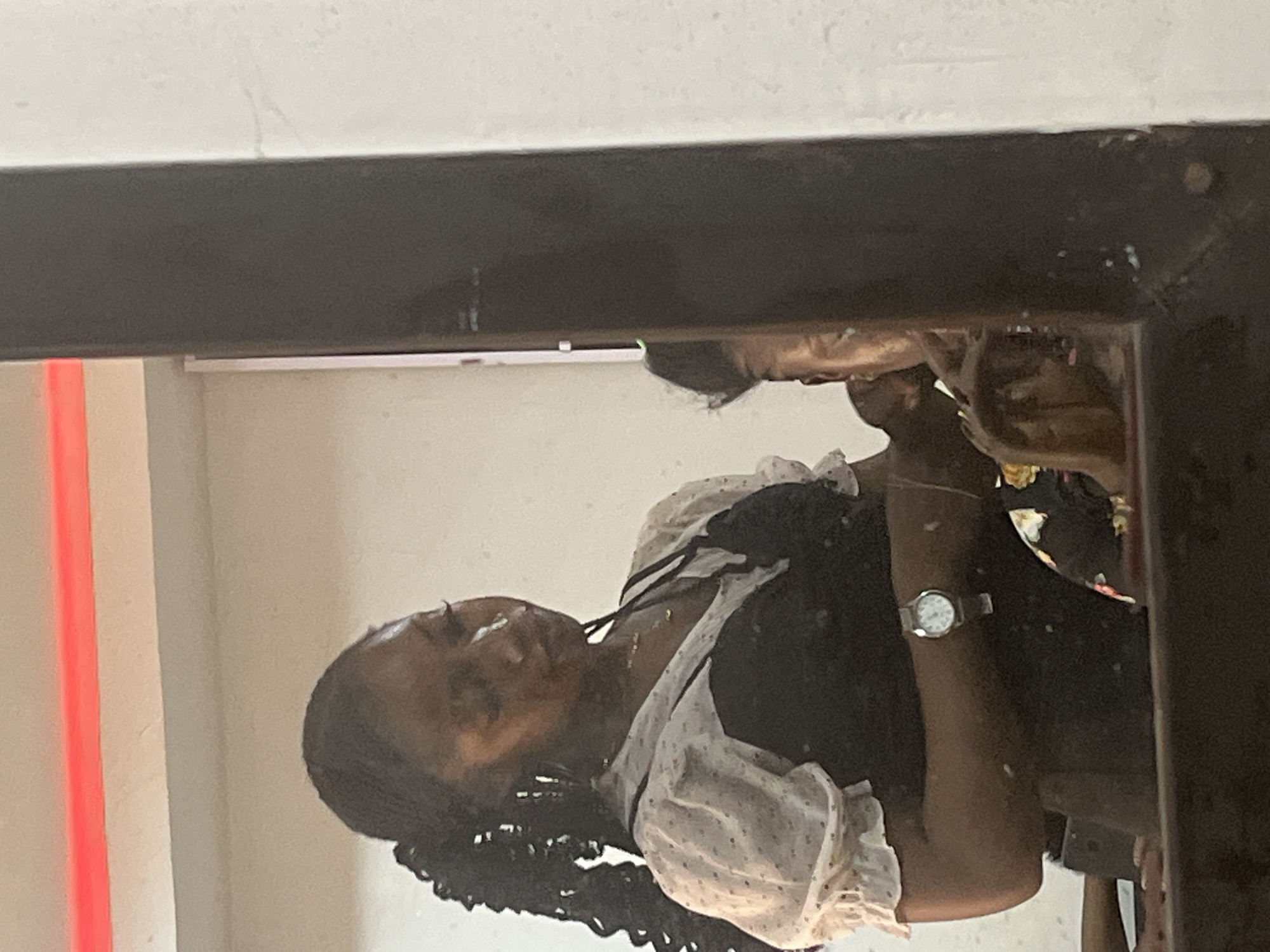 The image depicts an indoor scene, likely a salon or private grooming space, within Abuja, Nigeria, captured as a reflection in a dark-framed mirror. A female individual with a dark complexion is centrally featured in the reflection. She has long, dark braided hair styled to the left side of her head. Her attire consists of a white, short-sleeved blouse with small dark polka dots, worn beneath a dark, sleeveless top. A thin, gold-colored necklace is visible around her neck, and a silver-toned wristwatch with a white face is on her left wrist. Her head is tilted forward, with her eyes cast downward and partially obscured, indicating focus on a task. Her hands are positioned near the head of a second individual, whose partial reflection is visible in the lower-right quadrant of the mirror, suggesting she is attending to or styling this person. The second individual's clothing exhibits a multi-colored floral pattern with discernible yellow, orange, and black hues. The background features a light-colored wall, and a prominent horizontal red light strip extends across the top of the reflected scene. The dark frame of the mirror is visible along the right and bottom edges of the image.