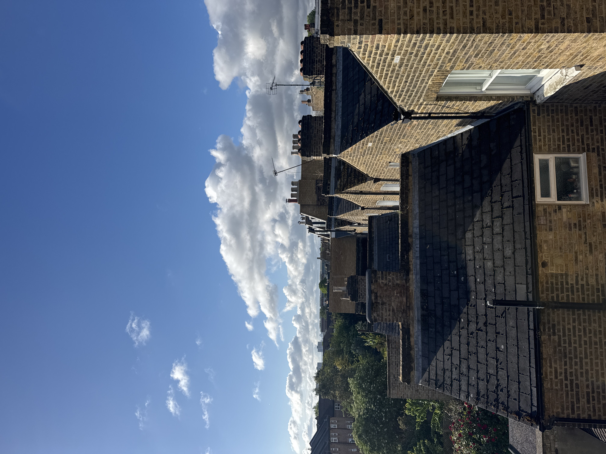 The image depicts a view over a residential area in London, United Kingdom. The perspective is from an elevated position, likely a window or rooftop, looking towards the horizon. The foreground is dominated by terraced houses with brick facades and slate or tiled roofs. Numerous chimneys are visible, many topped with traditional chimney pots, and television antennas are present on some roofs. Windows, some with white frames, punctuate the brickwork.
Beyond the rooftops, a line of trees indicates a park or green space. Further in the distance, low-rise buildings are visible, suggesting a residential area extending towards the horizon.
The sky occupies a significant portion of the frame, featuring a bright blue hue with scattered cumulus clouds. The lighting suggests a sunny day.