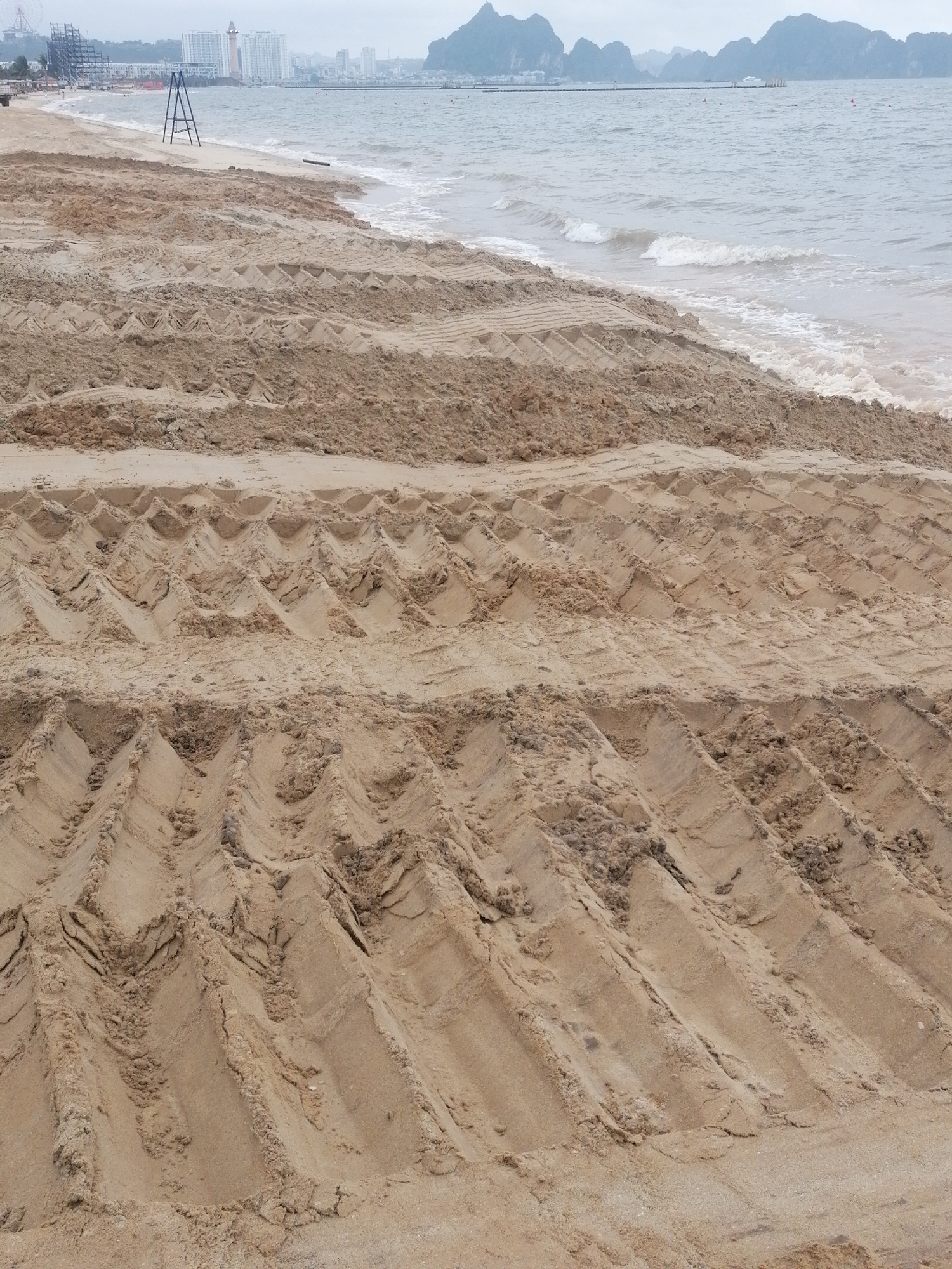 This wide-angle shot captures a coastal scene in Bai Chay Ward, Vietnam, under an overcast sky. The immediate foreground and midground are dominated by light brown sand, heavily marked with distinct, repeating V-shaped or zigzag patterns, indicating recent activity by heavy machinery, likely for beach grooming or sand replenishment. The sand is uneven, showing fresh disturbances.
To the right, the ocean stretches out with small, gentle waves breaking near the shore. The water appears a muted grey-blue. In the mid-distance on the left side of the beach, a tall, blue A-frame structure stands near the water's edge.
The background features a blend of natural and urban elements. On the left, a modern city skyline with multiple high-rise buildings and some construction is visible along the coast. Further into the distance, particularly on the right, characteristic dark, jagged limestone karsts rise dramatically from the water, suggesting the proximity of a landscape like Ha Long Bay. Several buoys are scattered in the water between the shore and the distant karsts.
The overall impression is one of a beach undergoing maintenance or development, contrasting the natural beauty of the karsts with urban expansion and human intervention on the coastline. There are no visible people or discernible text in the image. The soft, diffused lighting suggests an overcast day.