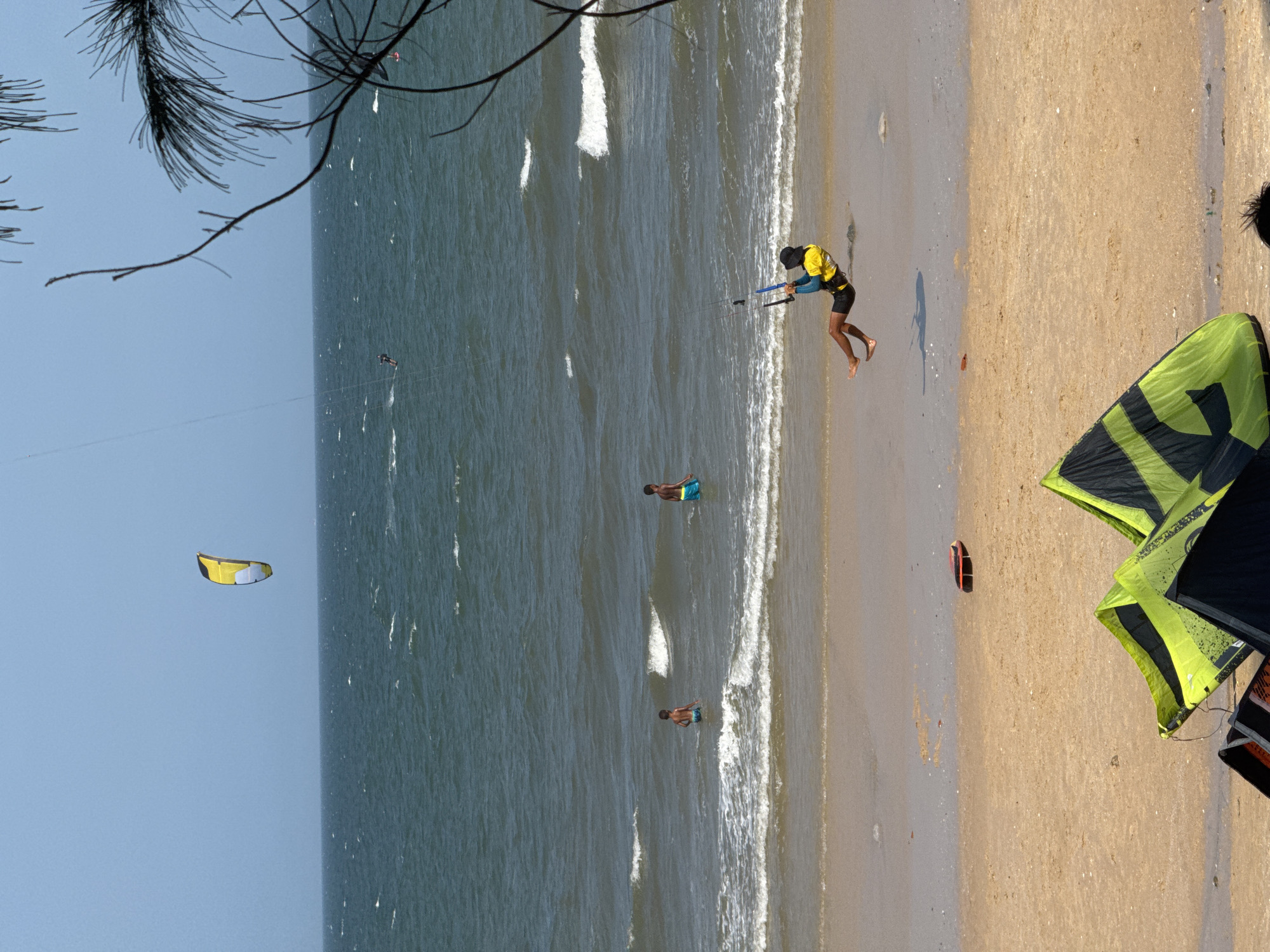 The image captures a lively scene at Mũi Né Beach in Phan Thiết, Vietnam, a popular destination for kitesurfing. The bright, sunny day is characterized by a wide expanse of blue sky meeting a choppy, dark blue ocean, with moderate waves crashing onto a sandy shore.
In the foreground, several kitesurfing kites are laid out on the sand, their vibrant lime green and black patterns stark against the beige beach. Further back, a kitesurfer is mid-action, suspended in the air over the water, demonstrating the sport's dynamic nature. This individual is wearing a yellow vest and black shorts, with a black hat providing shade. Their body is positioned dynamically as they are being pulled by the kite.
The middle ground of the image shows two young boys wading in the shallow ocean water, their dark skin contrasting with the white foam of the breaking waves. They appear to be enjoying the water. In the distance, other kitesurfers are visible on the water, suggesting a thriving kitesurfing community. One of these distant kites is distinctly yellow and blue, soaring high in the sky.
The background is dominated by the vast ocean and a clear, pale blue sky. The overall atmosphere is one of recreational activity and enjoyment of the coastal environment. A portion of a palm tree's fronds is visible in the upper right corner, framing the scene and adding a tropical element to the image. A red and yellow object, possibly a rescue buoy or a piece of equipment, rests on the sand near the water's edge.