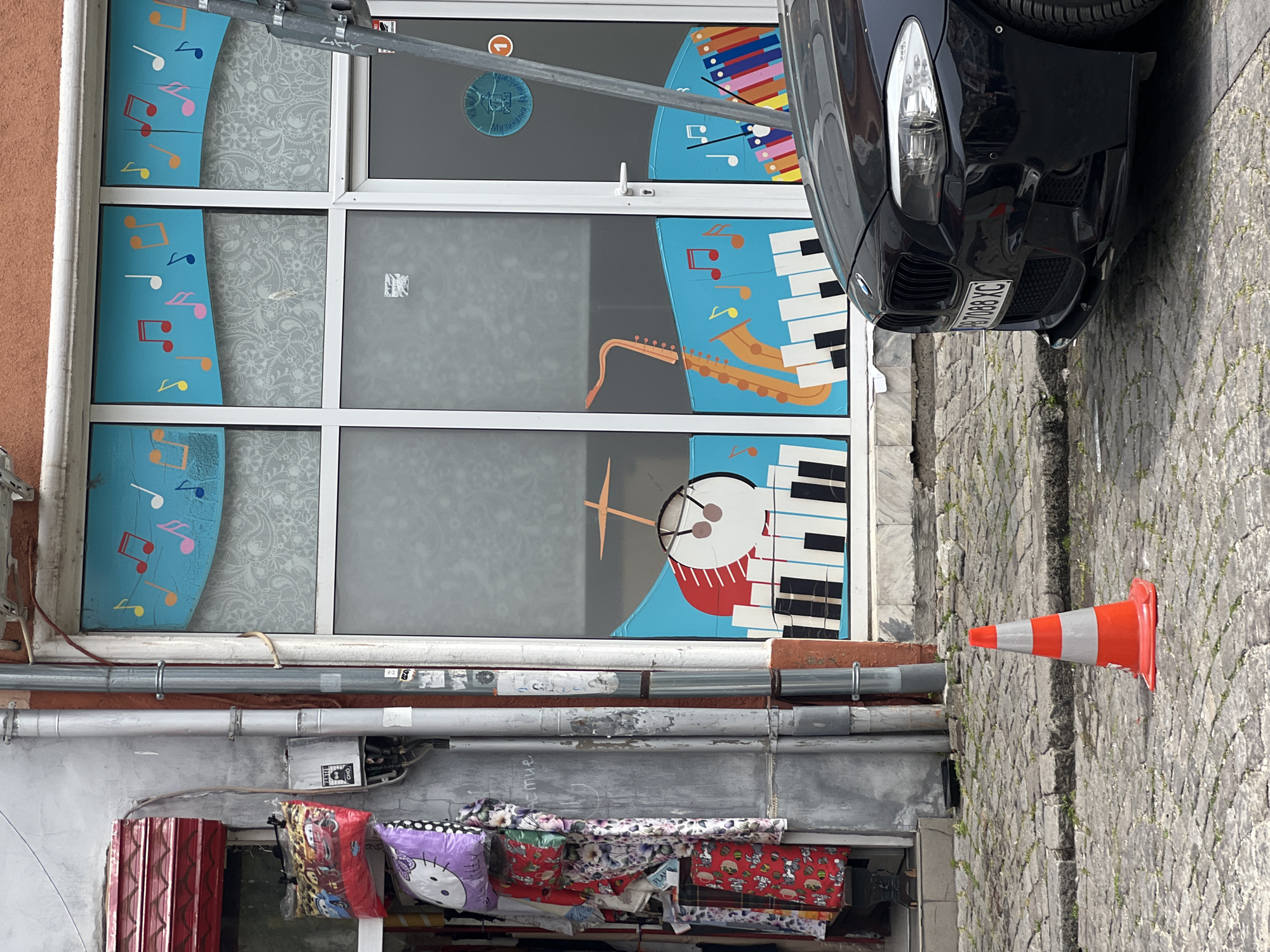 The scene shows the exterior of a building in Plovdiv, Bulgaria, where the front is decorated with musical instrument images on the windows, including a saxophone, drums, and keyboard. A black BMW car is parked next to the building, partially obstructing the view. An orange and white traffic cone stands on the sidewalk. Below the windows, there are some pipes and a window display of textiles.