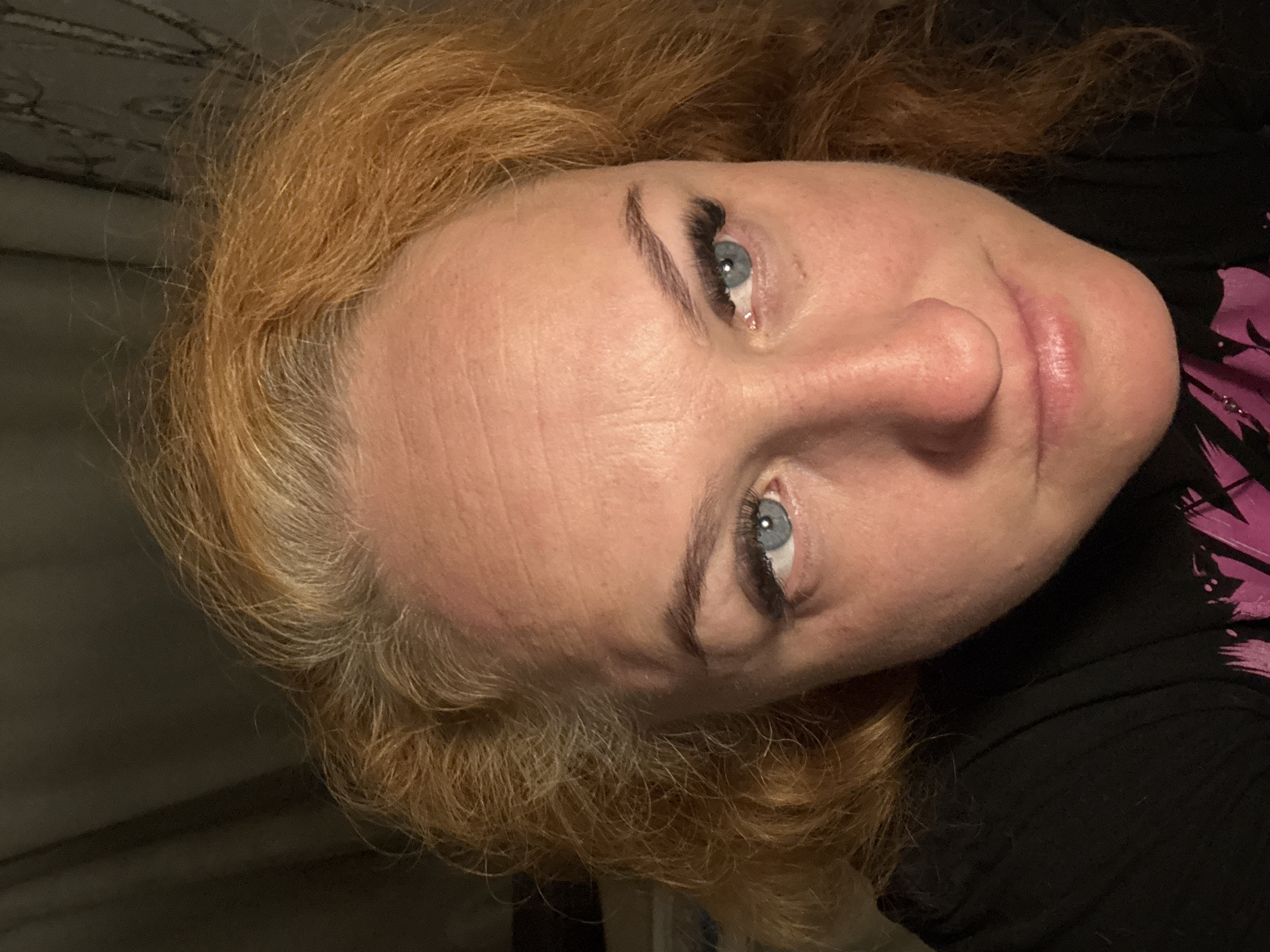 A close-up portrait shows a woman with light skin, blue eyes, and reddish-blonde hair. She is wearing what appears to be a black shirt with pink graphics on the right side. The woman is angled with her head tilted slightly. The background is a dark wall, possibly patterned. There is no information in the image that indicates the location in the city of Chaikovsky, Russia.