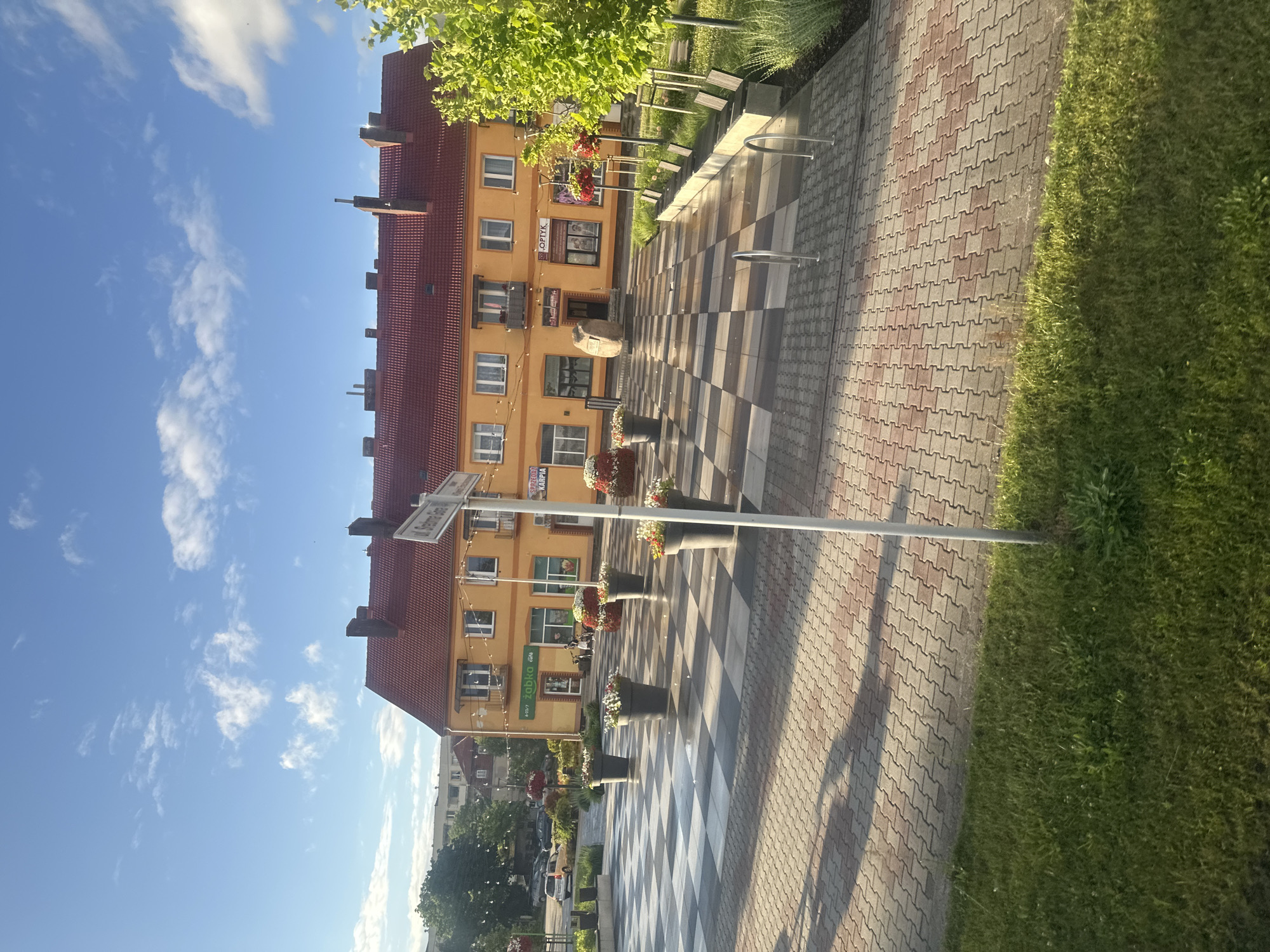 This is an exterior, eye-level, medium shot of a town square in Krosno Odrzańskie, Poland. The square features a checkered pavement pattern with large planters filled with flowers. A building with an orange facade and a red tile roof is visible in the background, with various businesses indicated by signs such as "zabka cafe," "SPRZEDAZ KARPIA," and "OPTYK." A street sign is also present. The sky is blue with scattered white clouds, and the sun is casting long shadows across the square. To the right, there is a row of benches and a small tree. In the foreground, a patch of green grass is visible. There are no people visible in the frame.