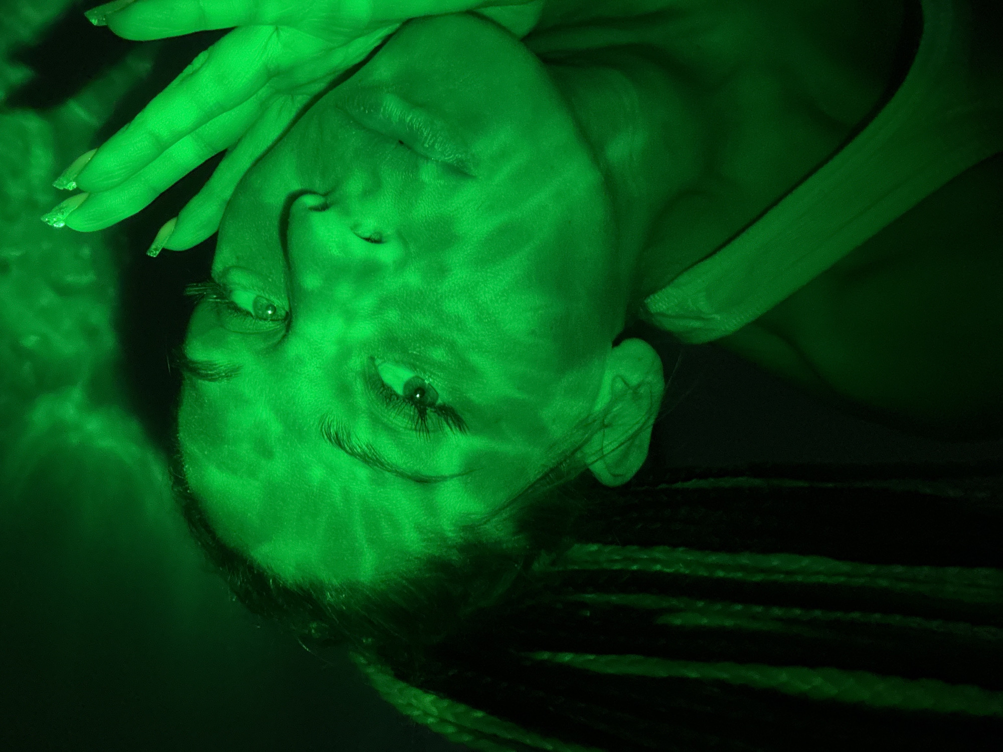 An artistic portrait photograph features a person, likely female, illuminated by a vivid green light in a dark setting. The subject's face is positioned vertically in the frame, with their head tilted slightly, and their gaze is directed towards the viewer. One hand, adorned with manicured nails that have a shimmery or glittered finish on the tips, rests on or near their forehead.
Distinct patterns of light and shadow, reminiscent of water reflections or projected textures, are cast across the person's face and hand, creating a dramatic and ethereal ambiance. The intense green illumination highlights facial features such as the eyes, eyelashes, and lips. Braided hair is visible towards the bottom right of the frame, largely blending into the dark background. A strap or piece of clothing is partially visible near the right side of the subject's neck or shoulder.
The setting appears to be dark, suggesting nighttime or an enclosed space where the artificial green light is the sole or dominant light source. The mood is mysterious and contemplative. No text is visible in the image. The scene is set in Rusanovo, Russia.