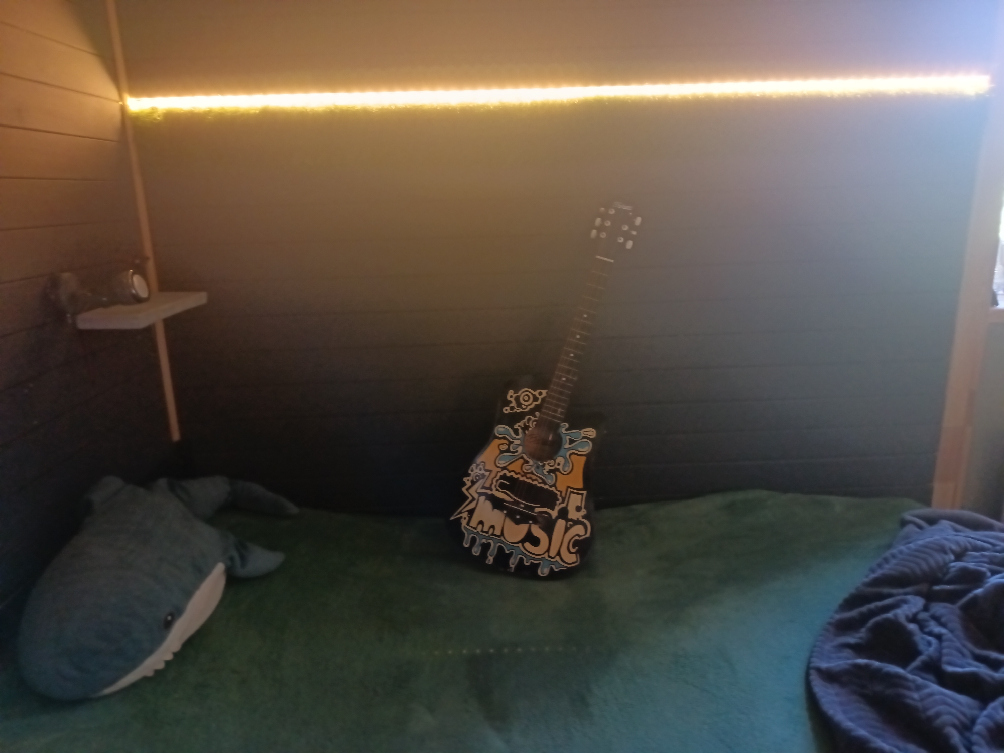 The image depicts an indoor scene, likely a room or a playhouse, featuring a bed covered with a green blanket. On the bed, a plush whale toy is positioned on the left side, and a small, decorated acoustic guitar is placed in the center. The guitar has colorful graffiti art on its body, including the word "MUSIC." In the background, the wall is paneled and a strip of warm yellow LED lights runs along the top. A small shelf is mounted on the left wall, holding a circular object, possibly a speaker or a small clock. The environment is dimly lit, with the LED lights providing a primary source of illumination. The overall impression is that of a cozy, personalized space. The location is not specified in the image or provided information.