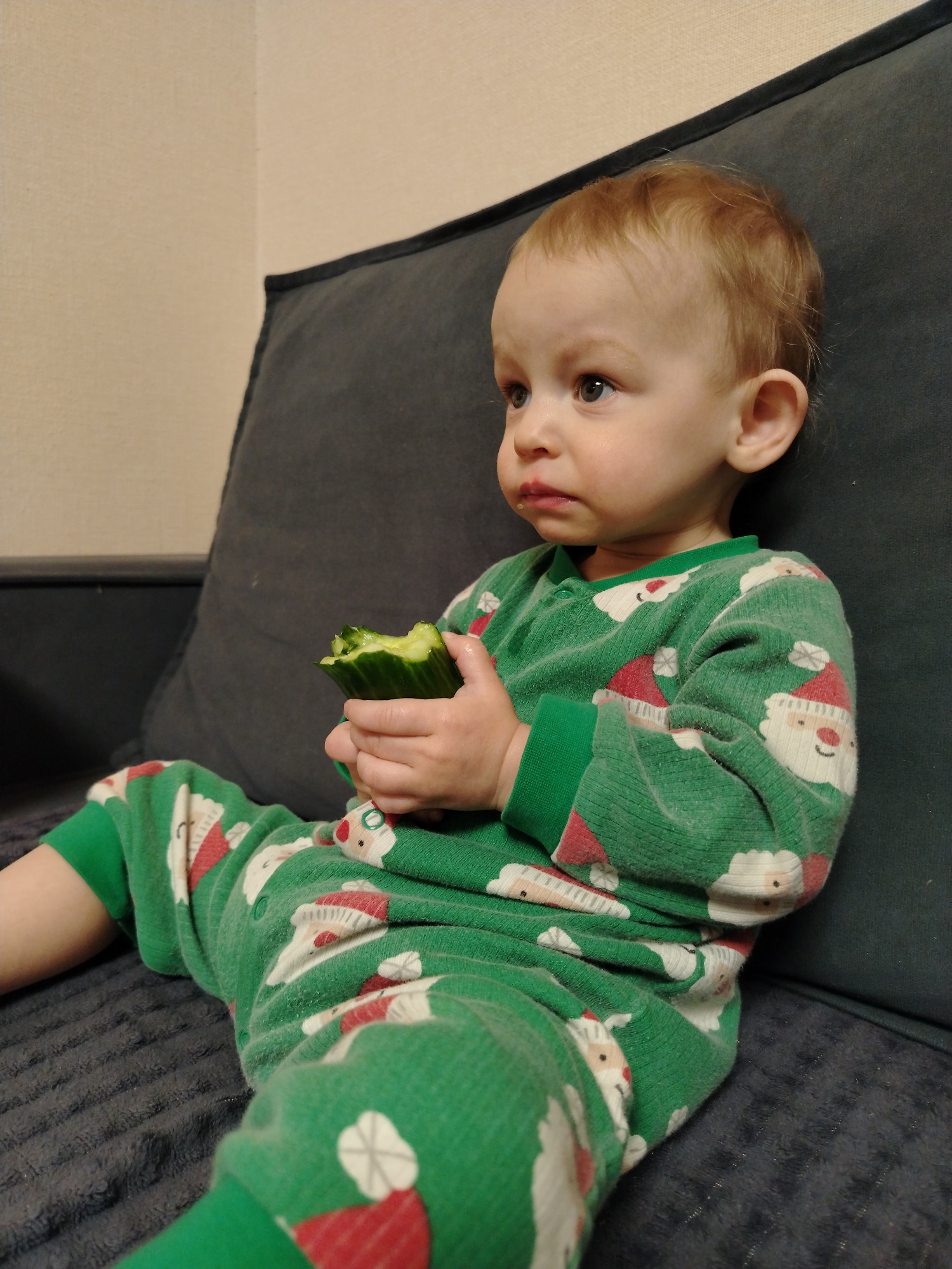 A young child wearing green pajamas with a Santa Claus pattern is sitting on a dark gray couch. The child is holding a partially eaten cucumber in their hands and looking off to the side with a slightly furrowed brow. The environment appears to be indoors, possibly a home. There is no explicit evidence in the image to confirm the location in Chaikovsky, Russia.