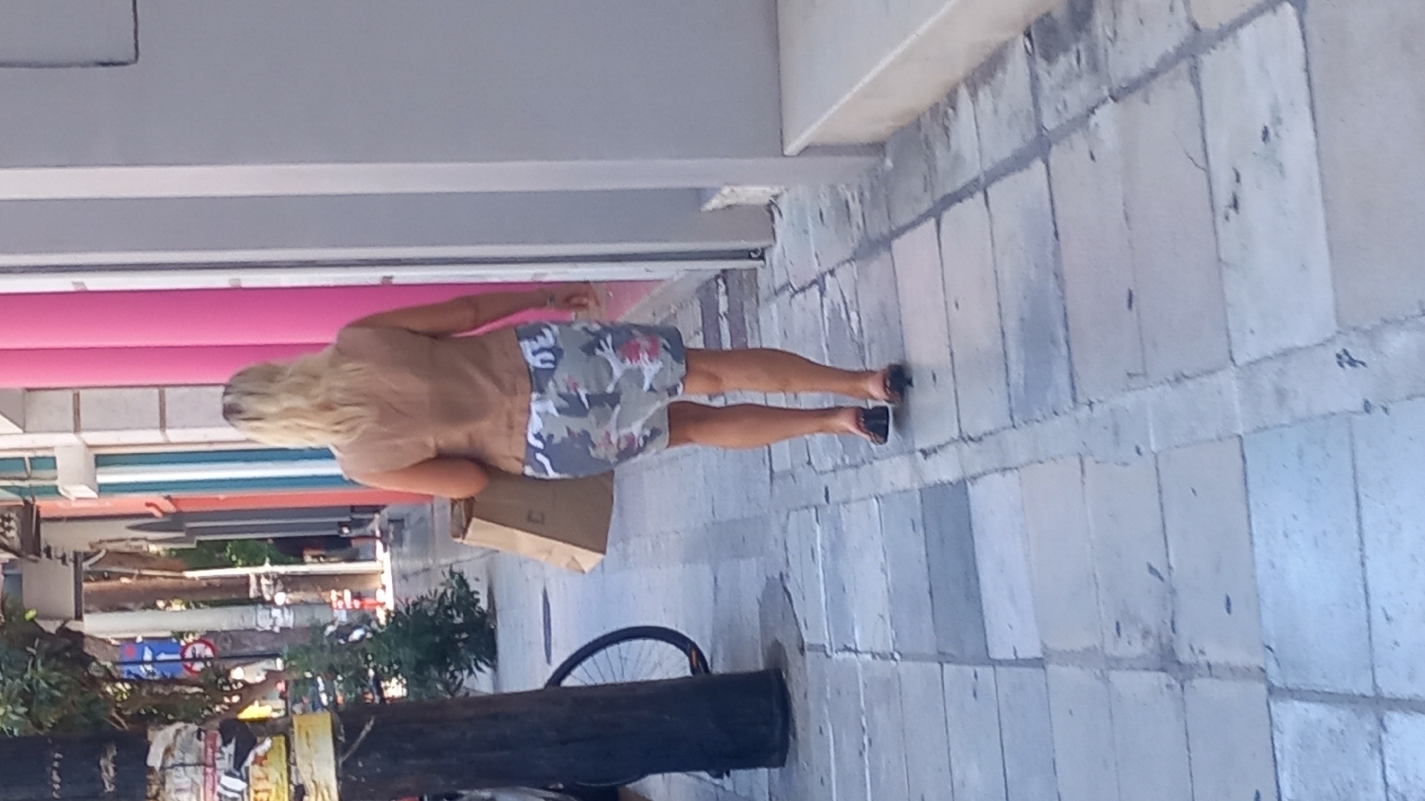 A blonde woman walks away from the camera on a cobblestone sidewalk in the city of Kos, Greece. She is wearing a tan sleeveless vest over a camouflage print skirt and black flip-flops. She carries a brown paper shopping bag in her left hand. The background shows a street lined with buildings featuring pastel-colored columns, some with awnings and signage. A dark tree trunk is visible to the left, with a bicycle wheel behind it. The sidewalk is made of large, grey rectangular stones.