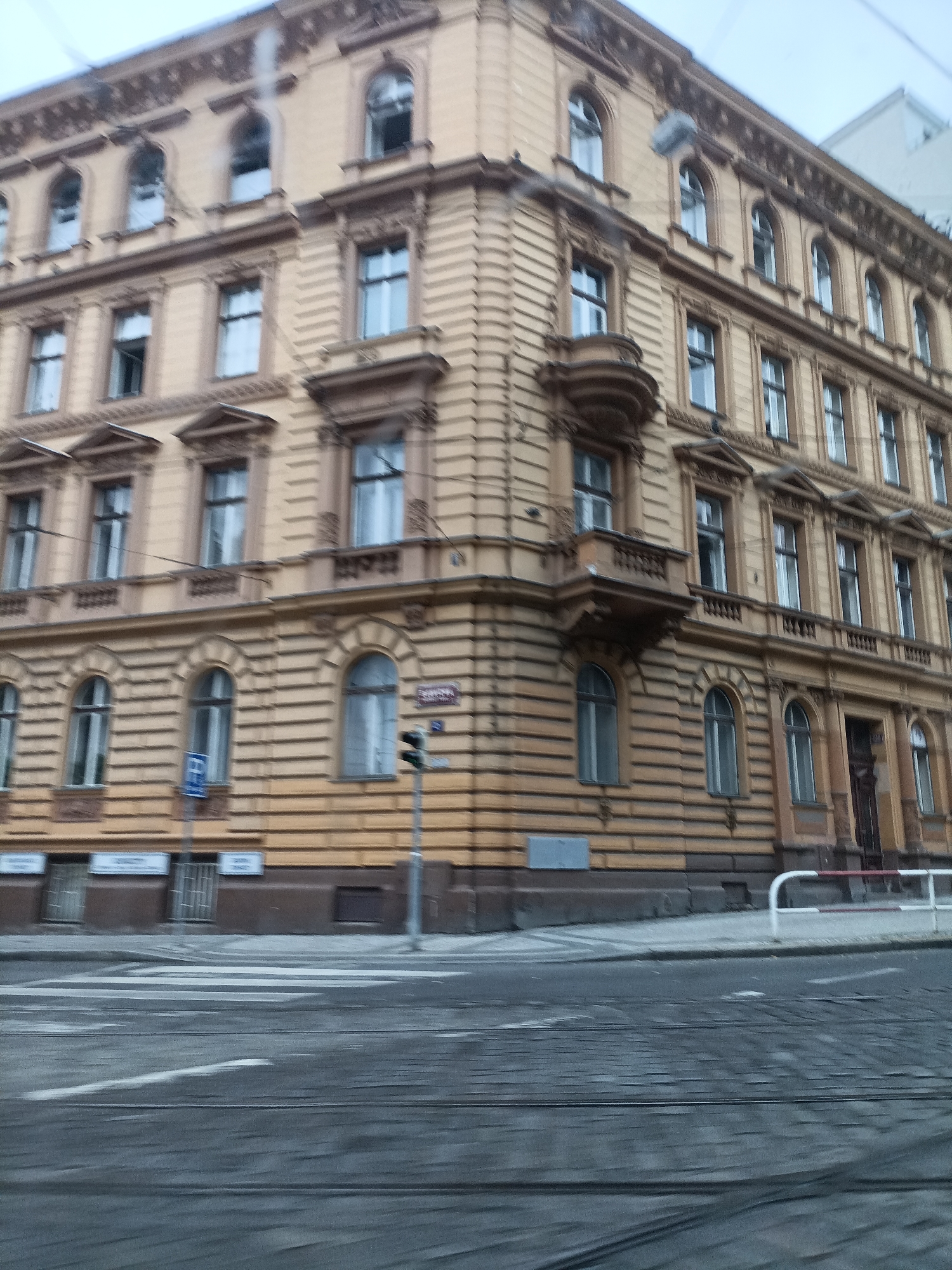 The image depicts a street scene in Prague, Czechia, on a cloudy day. The foreground is dominated by a paved street with tram tracks, suggesting a public transportation route. In the midground, the corner of a large, ornate, yellow building is visible. The building features multiple floors with numerous windows, some of which are arched, and decorative architectural elements. A traffic light and street signs are present on the sidewalk in front of the building. The background shows more of the building and a portion of a modern, light-colored building. The overall impression is of an urban environment with historical architecture. There are no people clearly visible in the image.