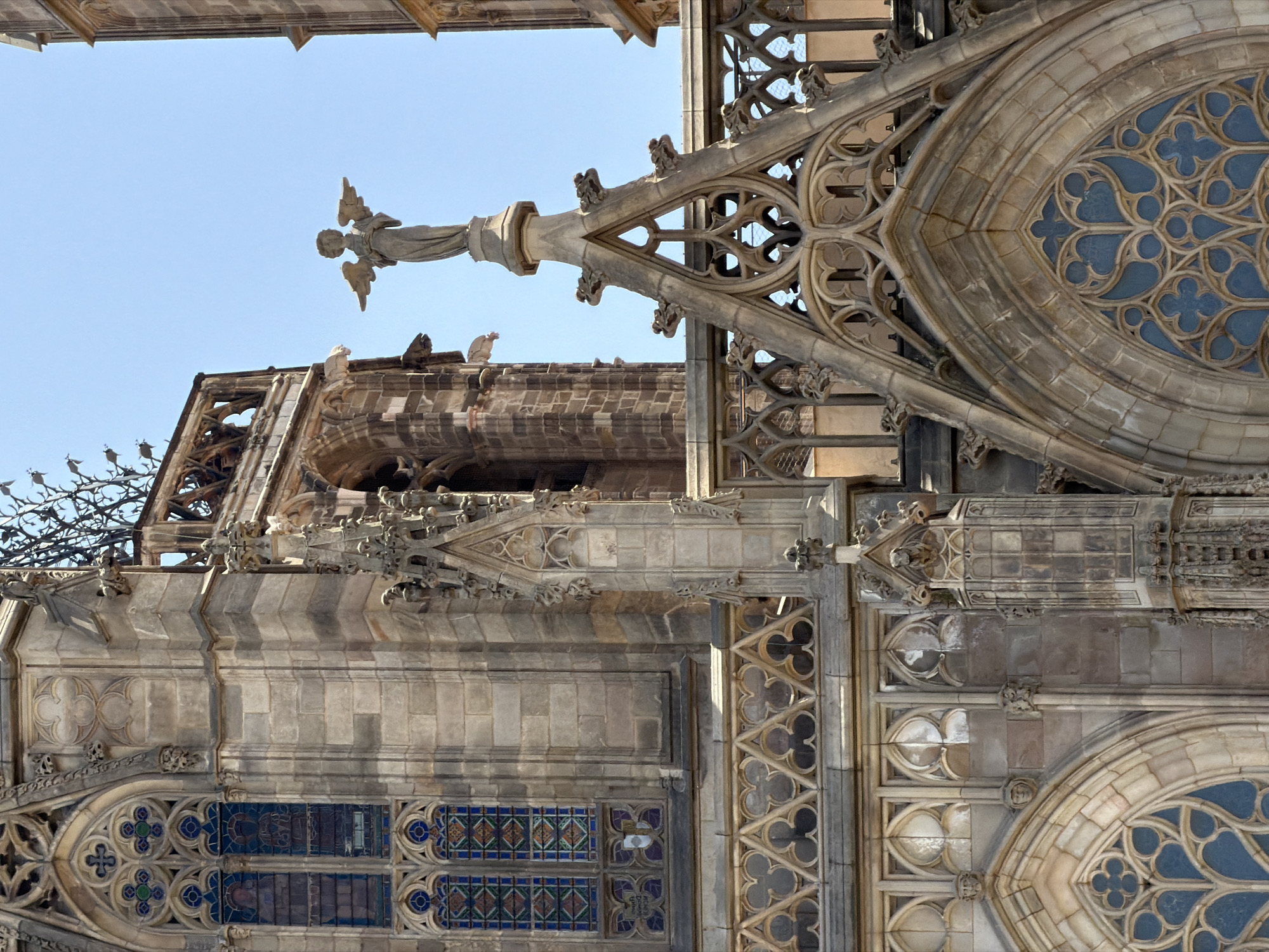 The image shows a close-up of the facade of the Barcelona Cathedral, located in the Gothic Quarter of Barcelona, Spain. The facade is constructed of weathered stone and features intricate architectural details typical of Gothic design.
Prominently displayed is a stained-glass window with a geometric pattern of blue and green panes. Below this window, a smaller section of stained glass depicts two figures. The building's facade is adorned with ornate carvings, including gargoyles and delicate tracery work along the balconies and window frames.
At the top of the structure, a stone statue of an angel with outstretched wings stands against a clear blue sky. The angel is positioned above a pointed architectural element, which is also decorated with carved details. The overall impression is one of grandeur and historical significance, showcasing the detailed craftsmanship of Gothic architecture.