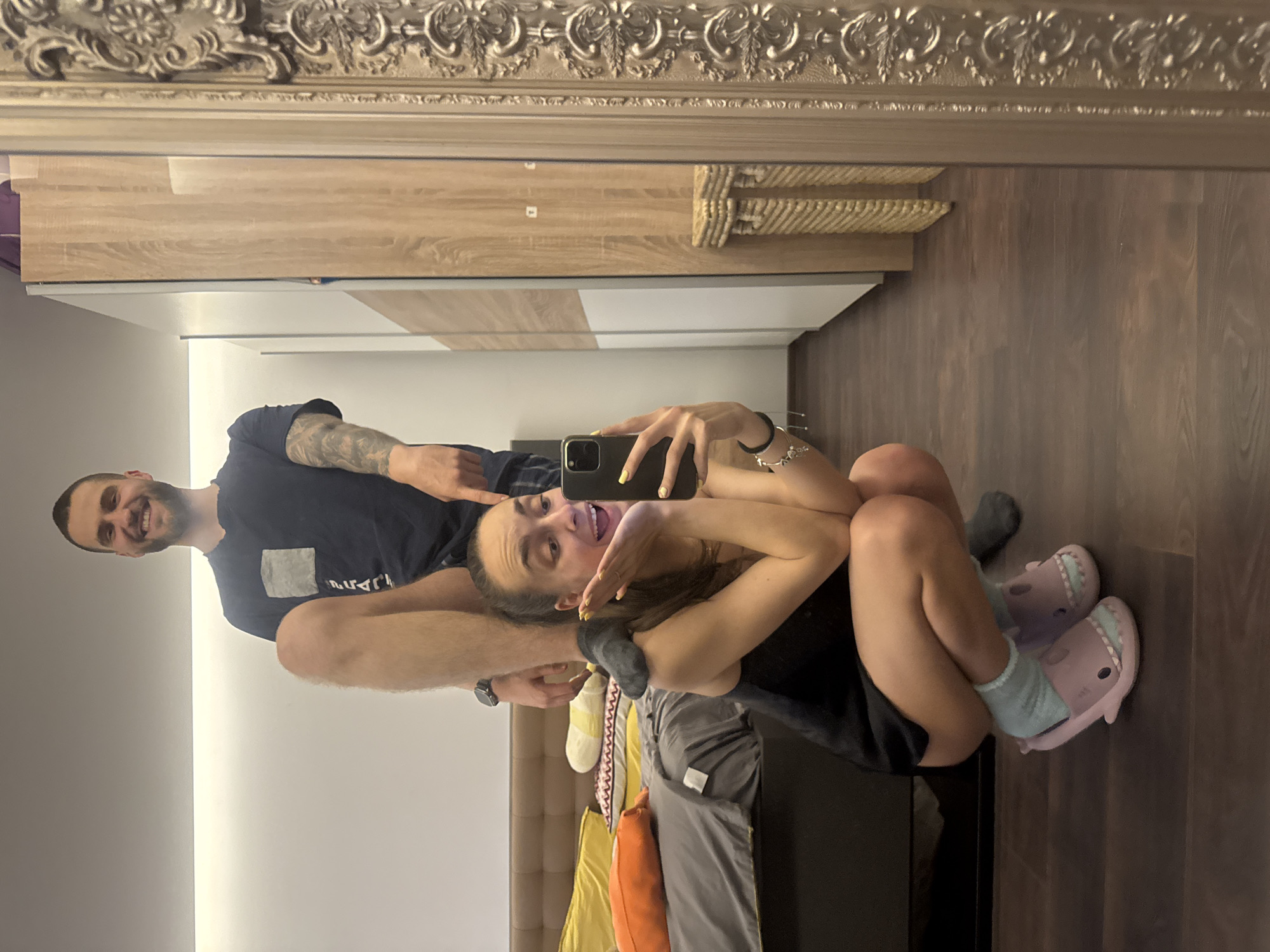 A man and woman are captured in a mirror selfie within an interior space. The man, sporting a beard and tattooed arm, is clad in a dark t-shirt. The woman, holding the smartphone, is wearing a dark top and pink shark-shaped slippers. The reflection shows a bed with colorful pillows, indicating a bedroom setting. A wardrobe with a light wood-tone finish is also visible. The location appears to be a private residence, possibly in the city of Sofia, Bulgaria, although there is no definitive evidence to confirm this.