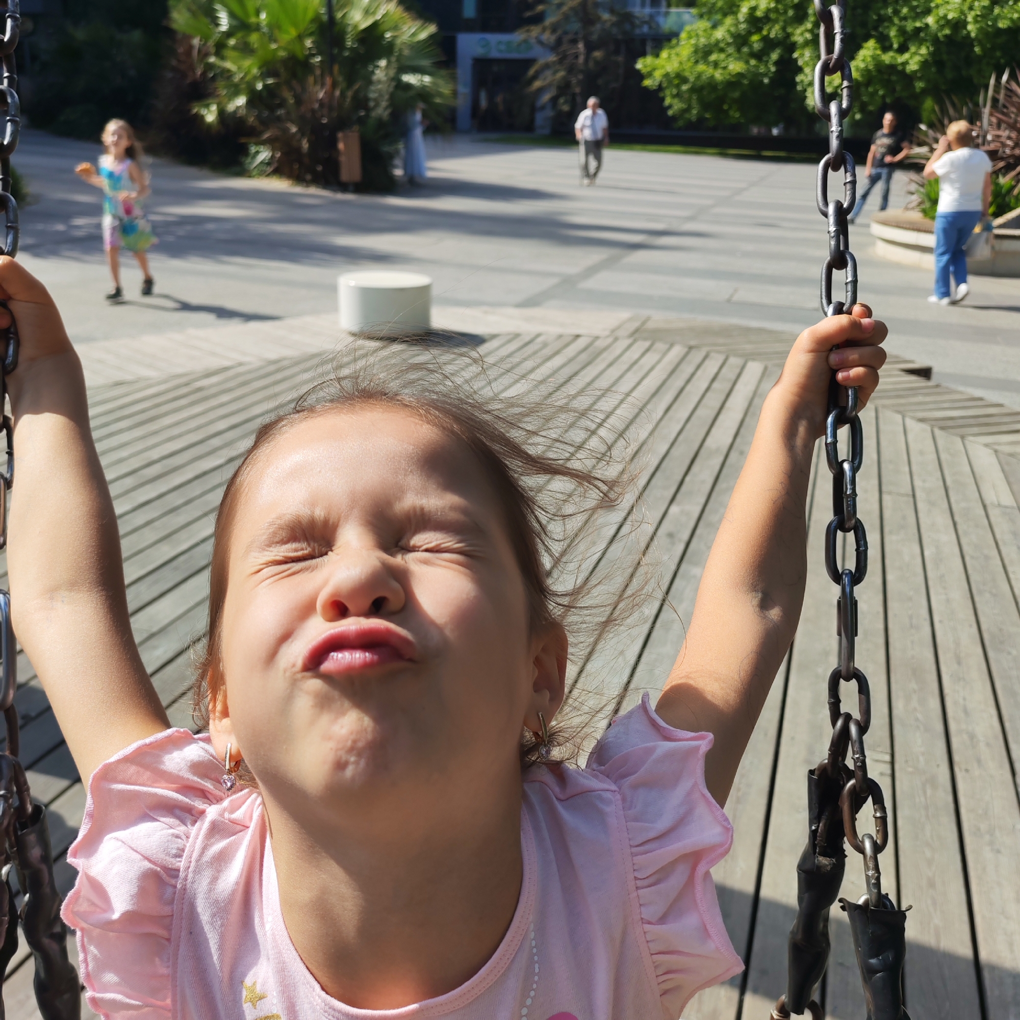 In the foreground of the image, a young girl is captured mid-swing on a swing set, her eyes squeezed shut and her lips pursed in a playful expression. She wears a pink t-shirt with ruffled sleeves and a heart-shaped earring. Her hand grips a heavy metal chain. The background shows a sunny outdoor public space with paved ground and wooden decking, suggesting a park or plaza. In the mid-ground, a woman in a white top and blue jeans walks away from the camera, and a man in a dark t-shirt is visible further back. In the distance, palm trees and other foliage indicate a warm climate. The setting appears to be an urban environment in the city of Yalta, Ukraine, given the lush vegetation and the architectural style of the building visible in the background.