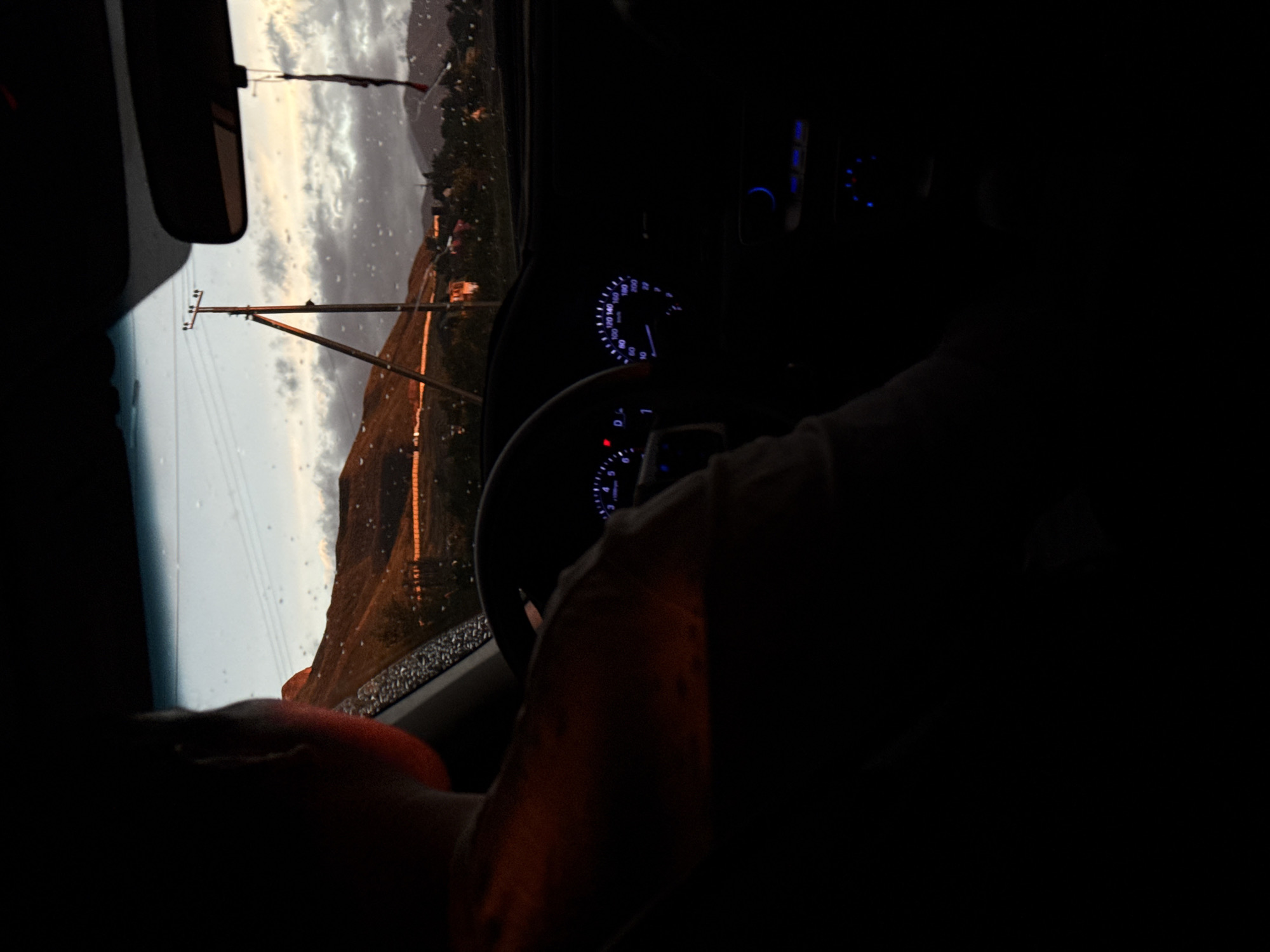 The image shows the interior of a car with a view of a mountain landscape through the windshield. It appears to be raining, as raindrops are visible on the glass. A utility pole is visible in the distance. The dashboard and steering wheel are partially visible, with the speedometer illuminated. A person's hands are on the steering wheel.