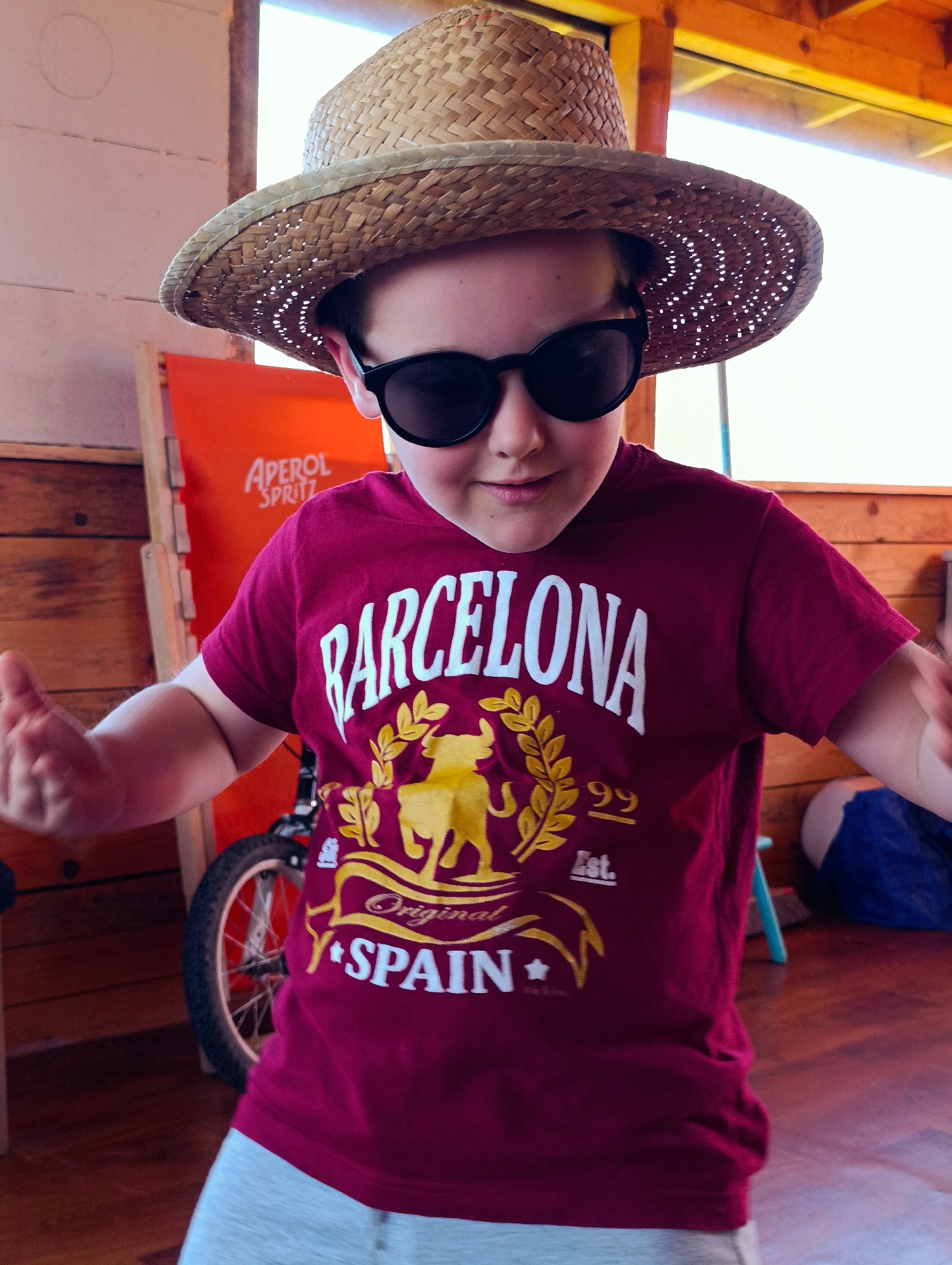 A child wears a straw hat and black sunglasses, a maroon t-shirt with "BARCELONA SPAIN" in white and gold lettering. The background includes a bicycle, an orange sign that says "APEROL SPRITZ", and wooden paneled walls. The location is Shiroko pole, Bulgaria.