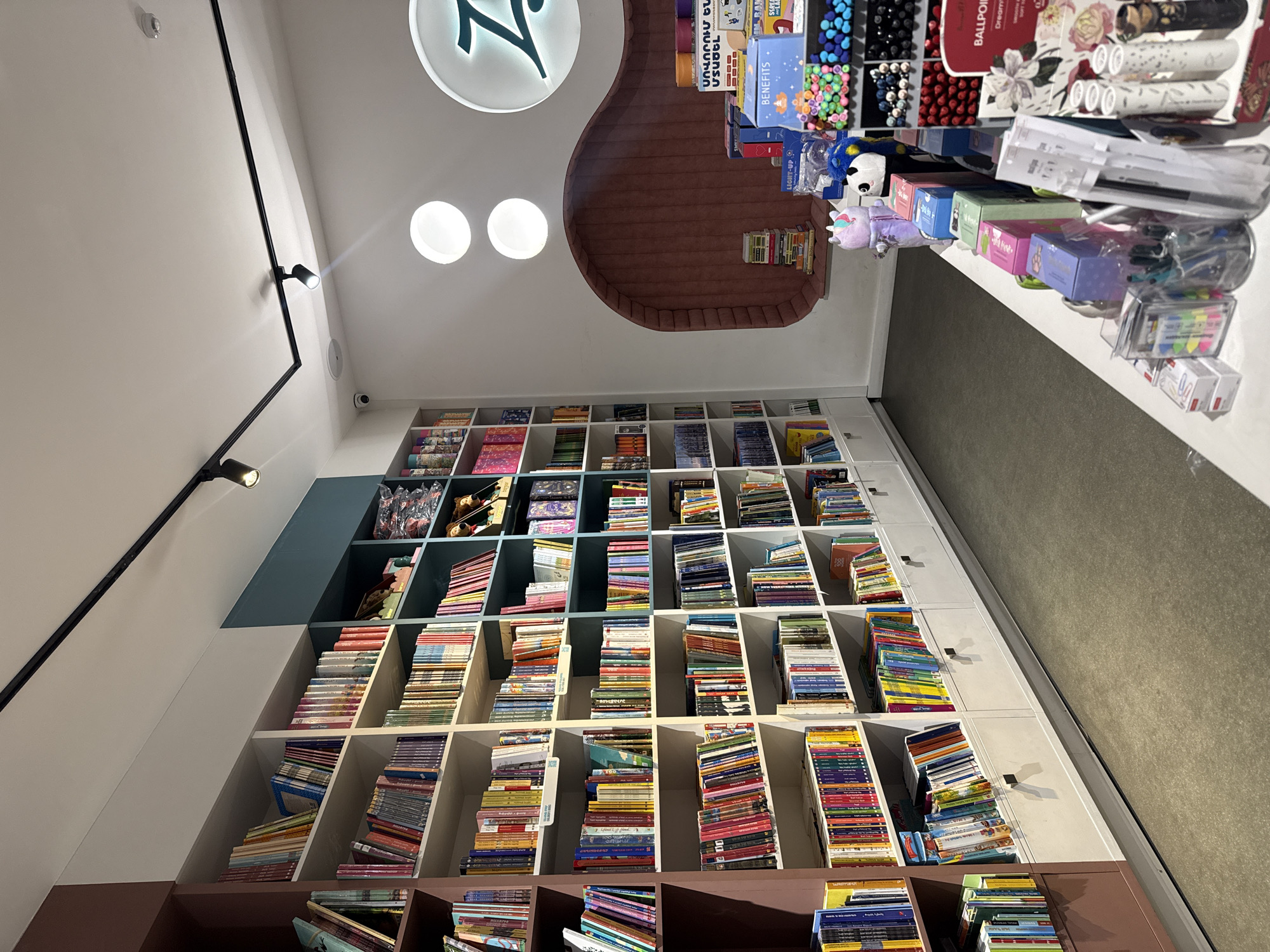 A vertically-oriented, upward-angled shot captures the interior of a store, likely a bookstore or a stationery shop, located in Yerevan, Armenia. The store features a tall, built-in bookshelf unit that extends almost to the ceiling, holding stacks of books arranged neatly within each section. A section of the top of the bookshelf is painted in a teal color. The bottom of the bookshelf is painted in a brown color.
The upper part of the image shows a section of the ceiling with circular lighting fixtures and a decorative element. At the top-right edge of the shot, merchandise such as pen sets, novelty pens, and other stationery items are positioned on a shelf.
The environment is clean and well-lit. The image does not show any people. The primary subjects are the bookshelves and the array of items on display, typical of a commercial retail setting.