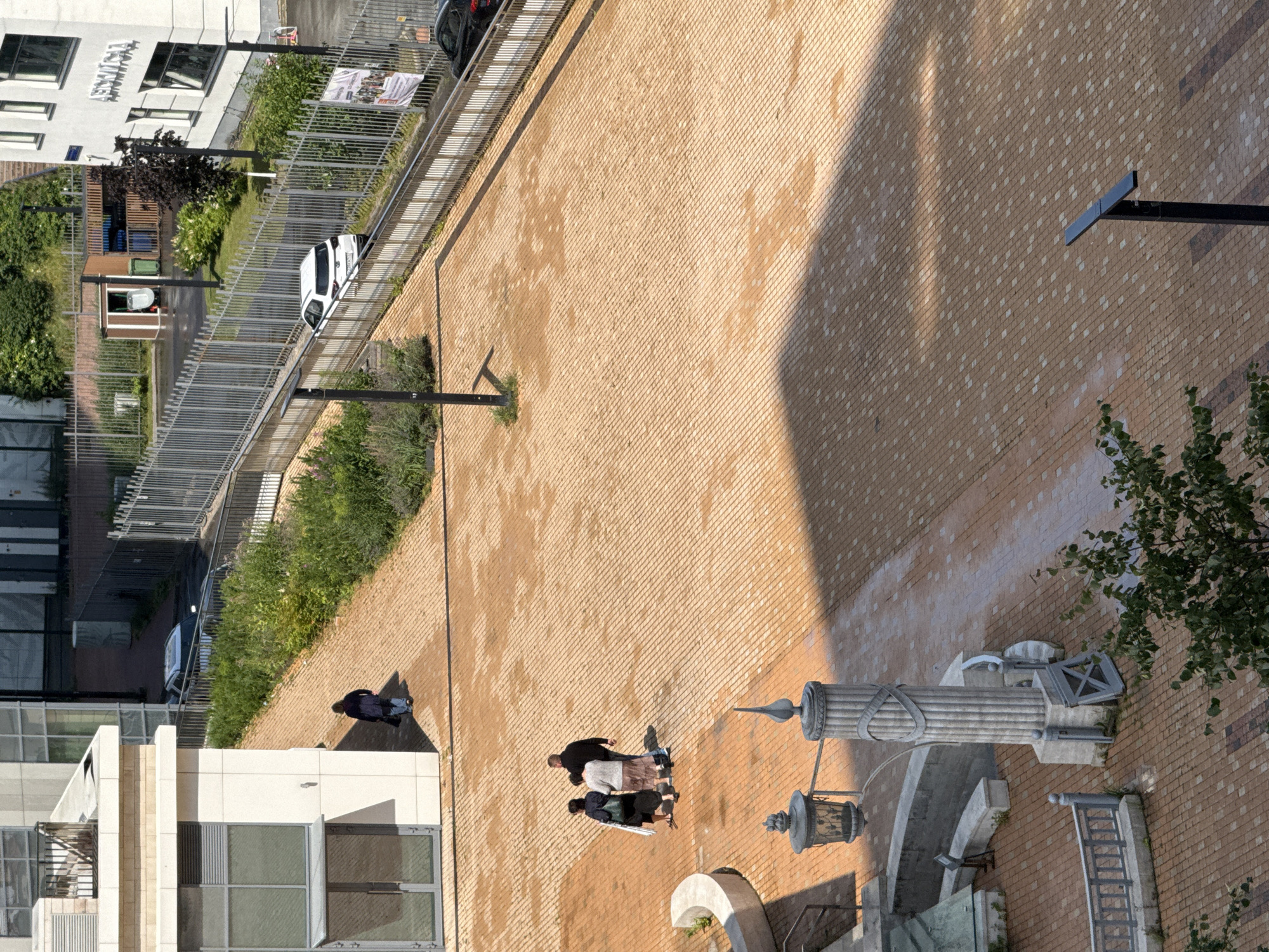 The image shows an aerial view of an outdoor paved area, possibly a courtyard or pedestrian zone, with an adjacent building and a fenced-off area beyond. The paving is made of small, reddish-brown bricks laid in a uniform pattern. In the foreground, a decorative stone pillar with fluting and some metallic embellishments stands prominently, next to a lamppost with an ornate metal shade. To the left, there's a curved stone bench.
Several people are visible. Near the center of the image, three individuals are walking together on the paved area. One person appears to be a man in a dark shirt, and two others are women in lighter-colored clothing. Further away, near the building on the left, a single person in a dark jacket and blue jeans walks away from the camera.
Beyond the paved area, there's a paved pathway with a metal railing, bordered by green bushes and grass. A white SUV is parked on this pathway. A metal fence runs along the perimeter of this area, separating it from a street or further complex where another car, black in color, is visible. A building with large windows and what appears to be signage in Russian (likely "Аптека" meaning "Pharmacy") is visible on the right. A small wooden structure with a green bin and a trash can is present in the fenced area. The scene appears to be urban, under daylight. Based on the signage and general architecture, the location is likely Moscow, Russia.