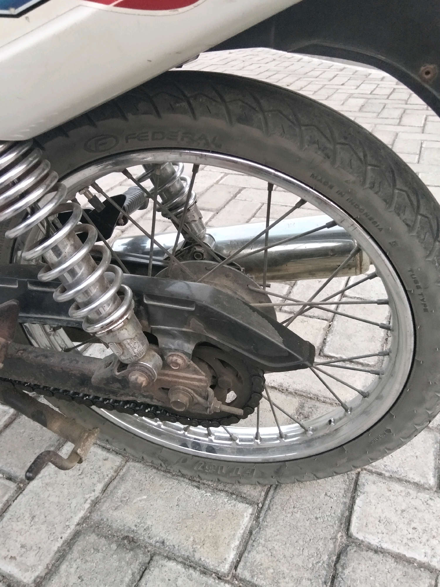 The image is a close-up, low-angle shot of the rear wheel and suspension of a motorcycle parked on a paved surface, likely a sidewalk or road, composed of grey interlocking bricks. The motorcycle is predominantly white with red and blue accents visible on the bodywork near the top left of the frame. The rear wheel is a spoked silver rim with a black tire. The tire bears the brand name "FEDERAL" and the inscription "MADE IN INDONESIA TUBE TYPE". The motorcycle's chain drive, rear brake mechanism, and a silver rear shock absorber with a coiled spring are clearly visible. A chrome exhaust pipe is also present behind the wheel. The ground is made of light grey, rectangular paving stones arranged in a staggered pattern. The overall condition of the motorcycle parts suggests some wear and tear, with dirt and grime present on the chain, brake components, and shock absorber. The scene appears to be static, with no people or activity depicted. The context of Jenangan, Indonesia, is a geographical identifier and does not provide visual information directly from the image.