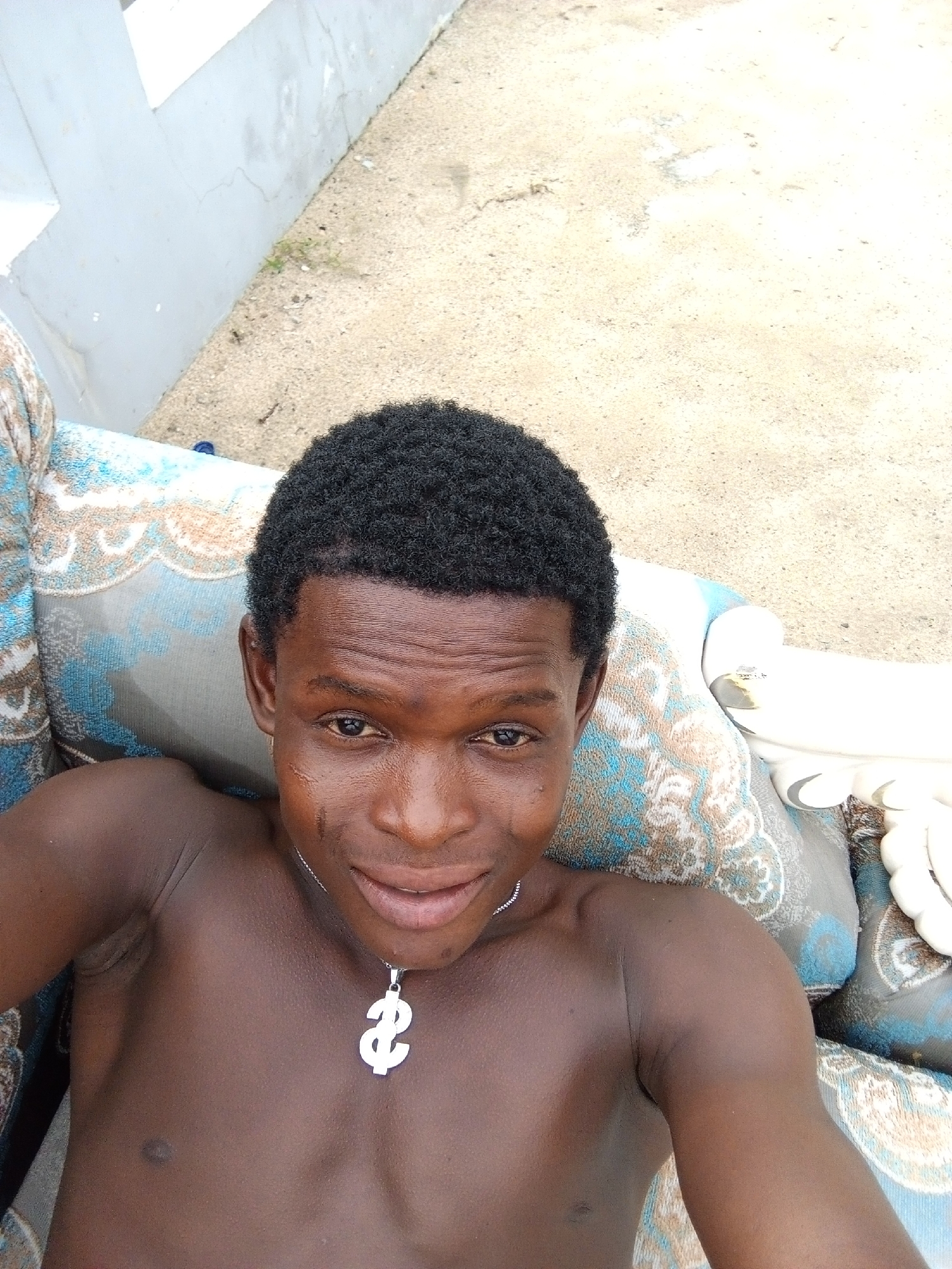 A medium-angle eye-level shot shows a shirtless dark-skinned man with close-cropped black hair and a silver dollar-sign pendant necklace. He is lying back on a blue and brown patterned upholstered couch. The man appears to be outside. In the background is an off-white painted wall and a sandy or concrete surface. It's difficult to place the specific location in Ado, Nigeria, based solely on the image.